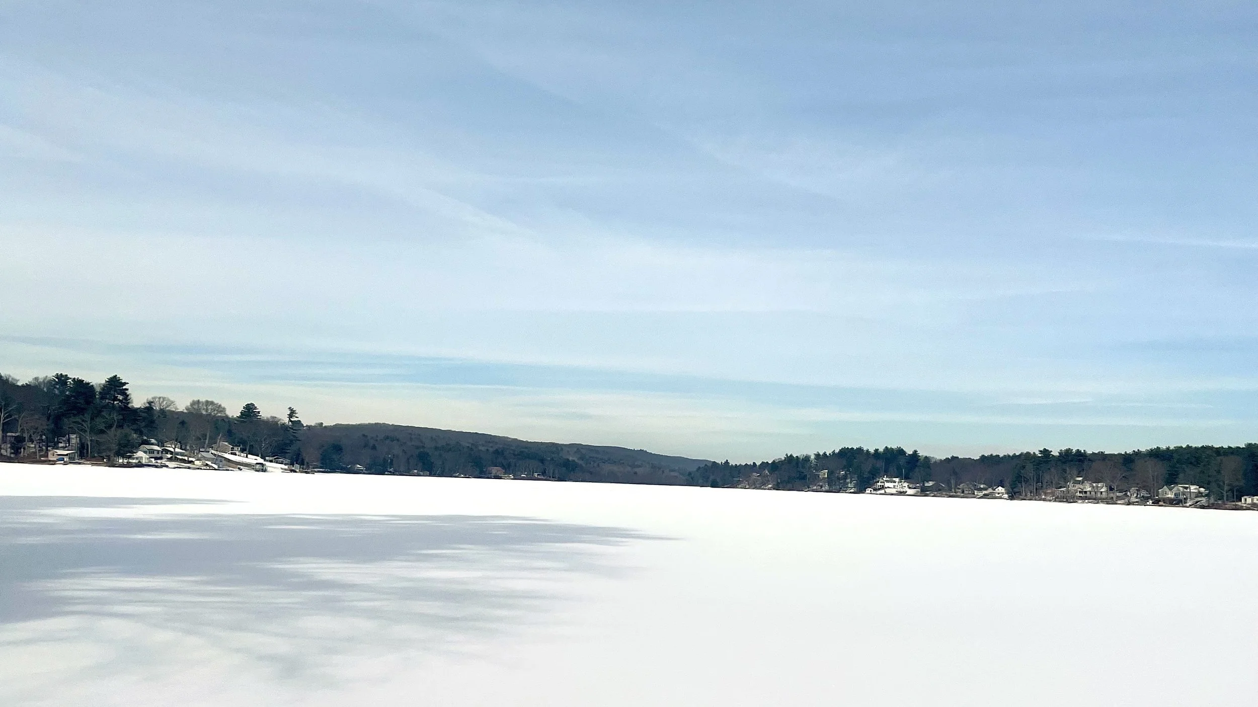 Snow-covered frozen lake with a distant shoreline lined with houses and trees under a partly cloudy sky.