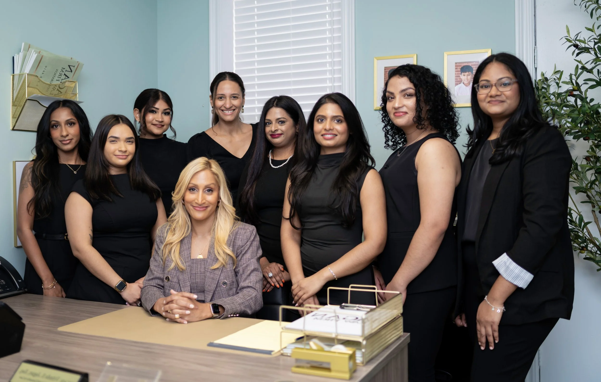 A group of ten professional women in business attire posing in an office behind a desk, with framed photos and plant in the background.