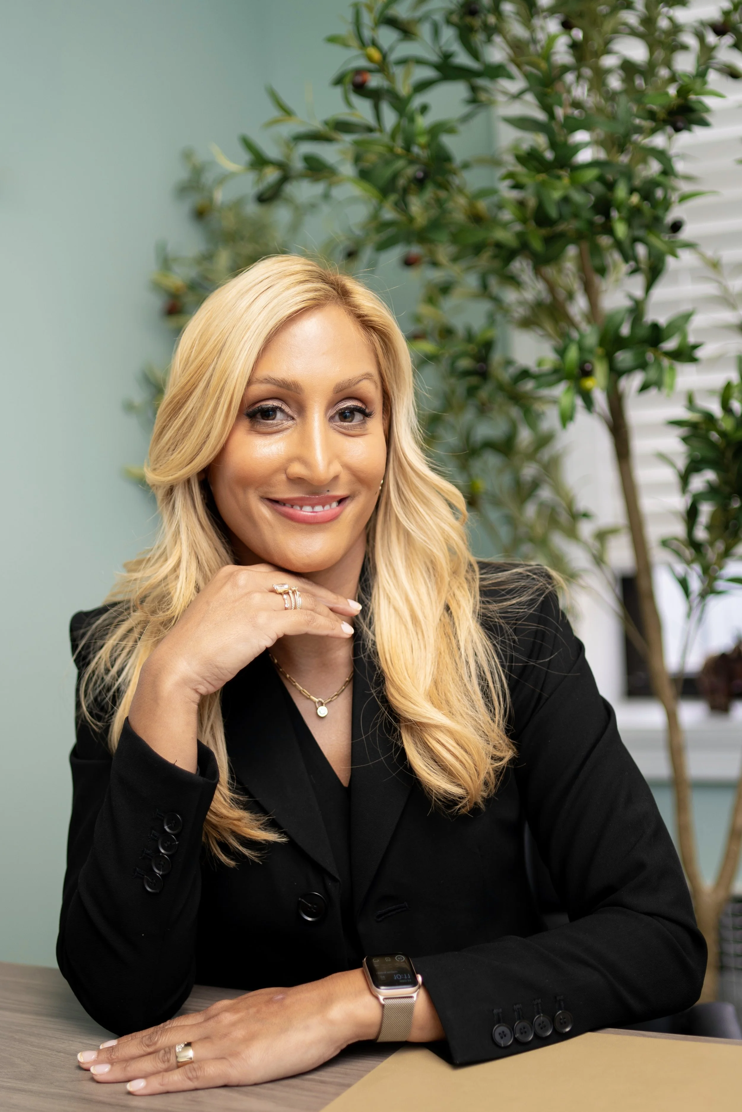 A blonde woman in a black blazer sitting at a desk with a smartwatch and jewelry, smiling at the camera, with plants and window blinds in the background.