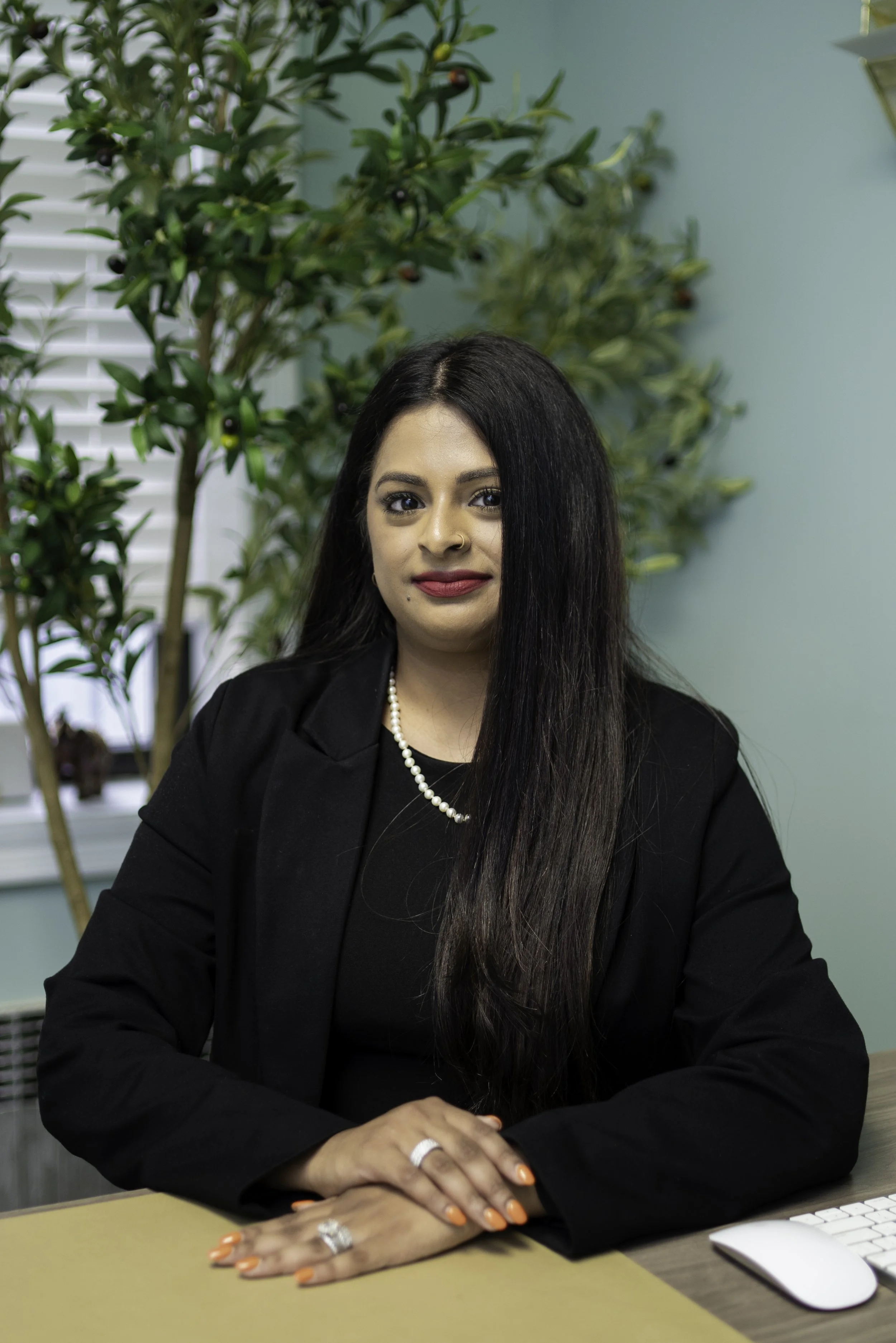 A woman with long black hair, wearing a black blazer, pearl necklace, and jewelry, sitting at a desk with a computer mouse, in front of a green wall with a plant background.