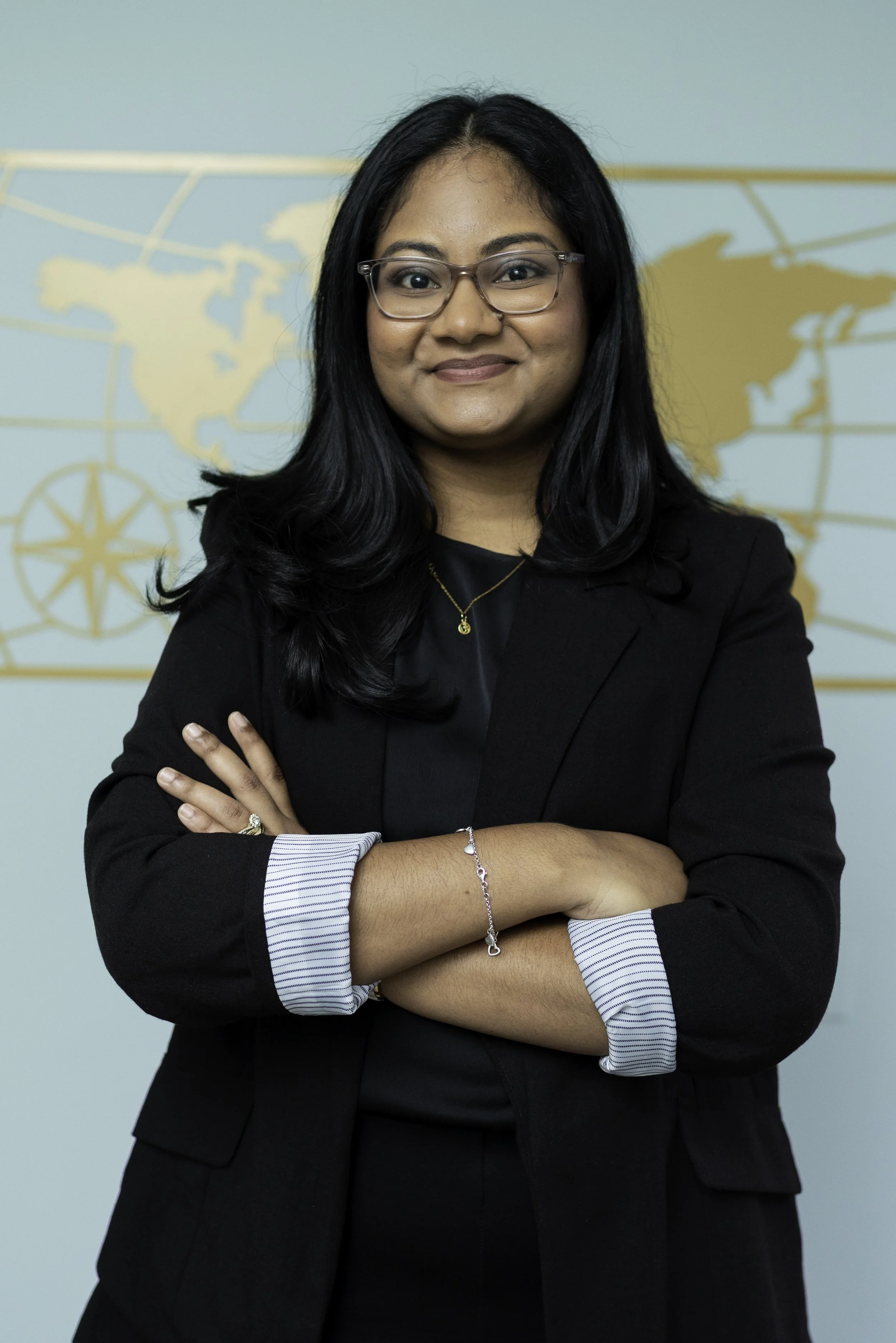 A woman with long black hair, glasses, and a black blazer crosses her arms and smiles at the camera. She is standing in front of a world map wall.
