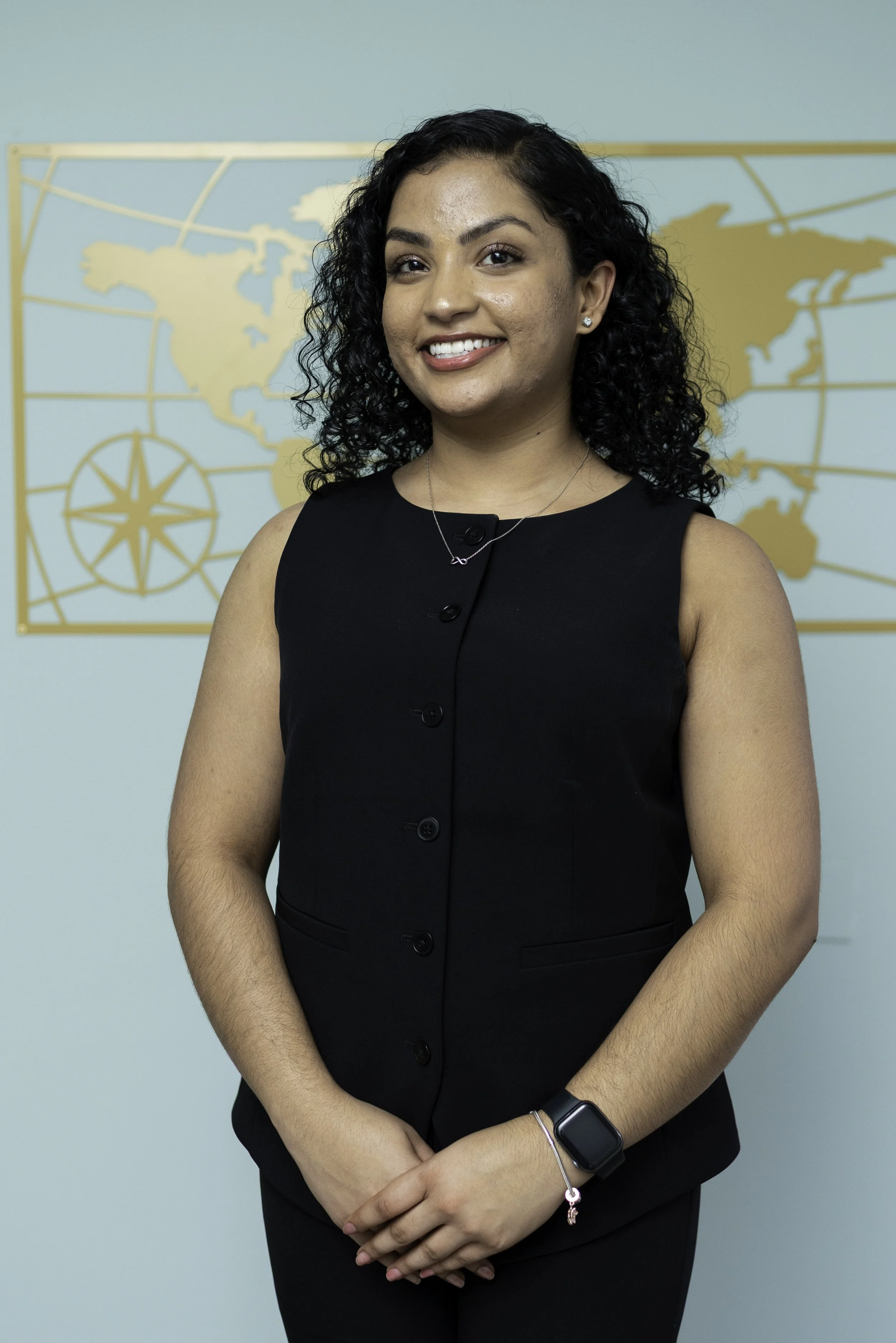 A woman with curly black hair smiling, wearing a black sleeveless top, standing in front of a world map poster.