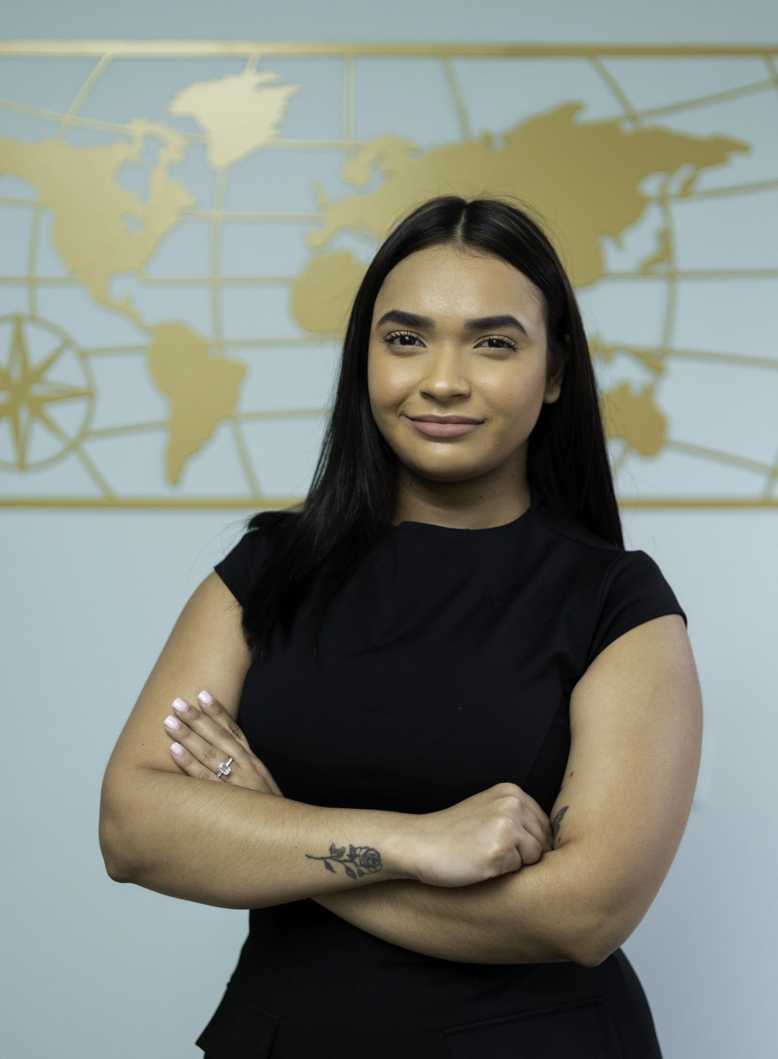 A woman with long black hair, wearing a black top, standing in front of a world map on a light blue wall, with her arms crossed and smiling.