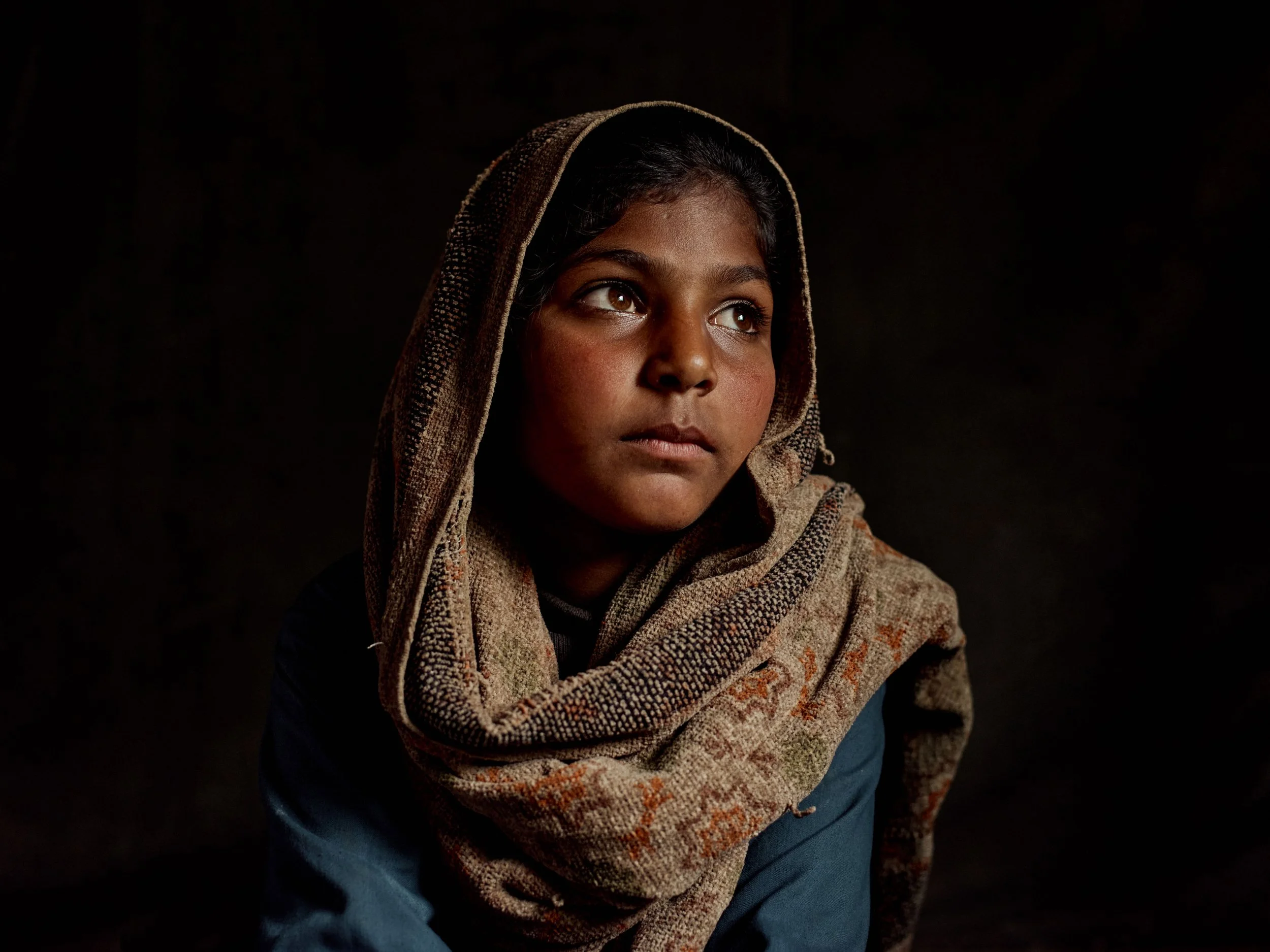 Portrait of a young girl with dark hair and brown skin, wearing a patterned headscarf and blue clothing, looking off to the side against a dark background.