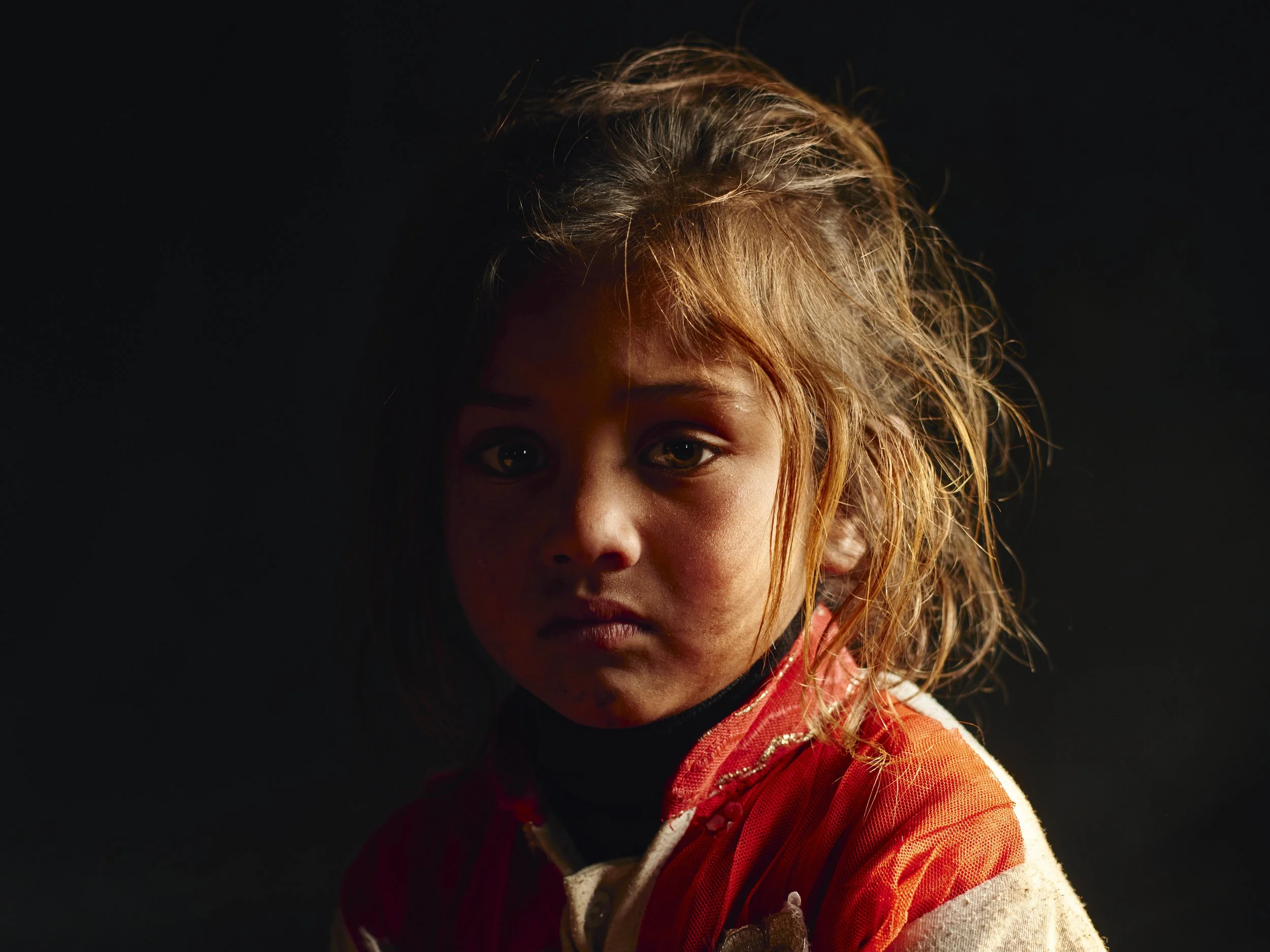 A young girl with messy hair and dirt on her face, wearing a red and white shirt, looking directly at the camera against a dark background.