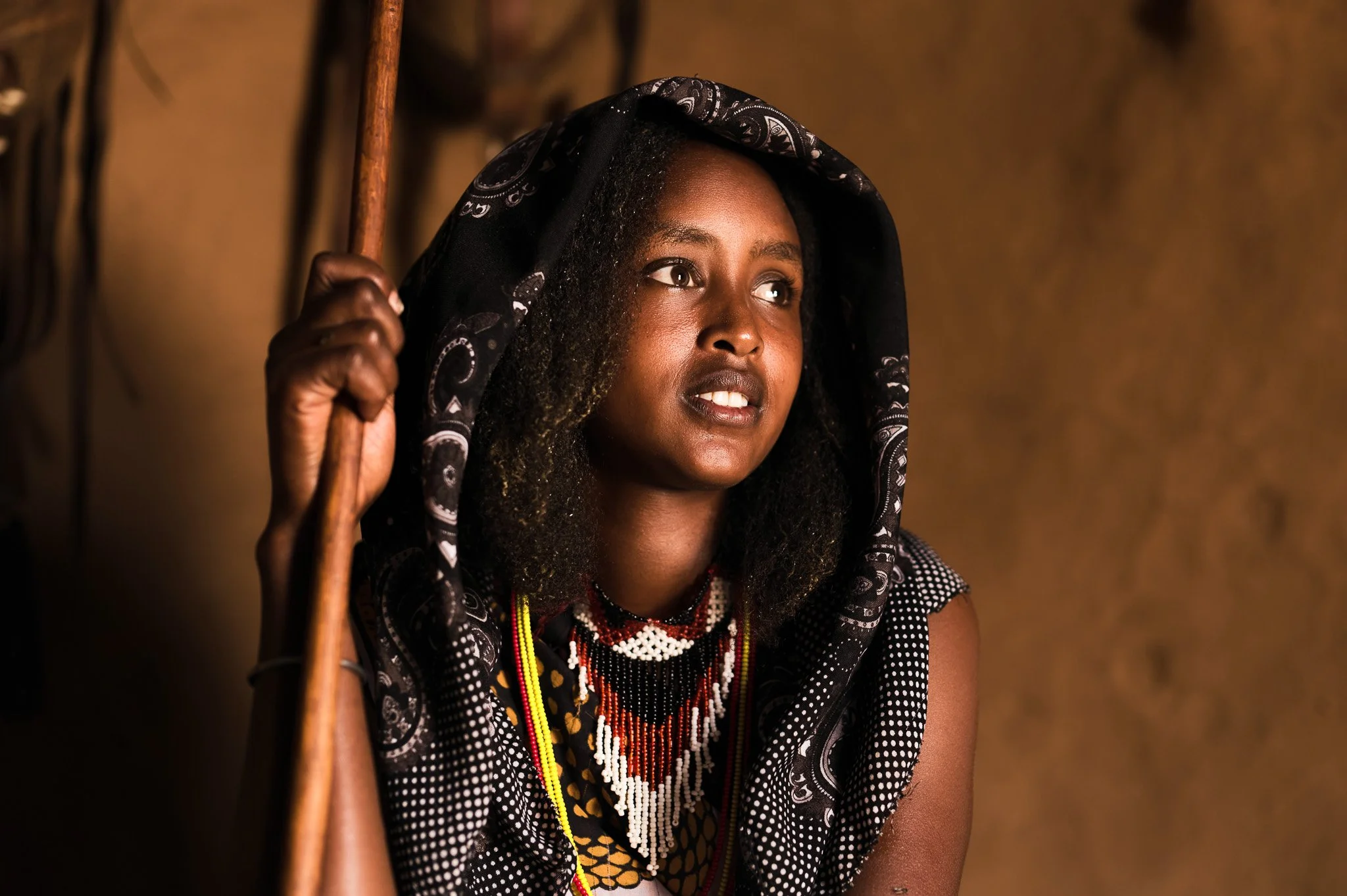 A woman with dark skin, wearing a black hooded cloth with white patterns and beaded jewelry, holding a stick, with a blurred brown background.