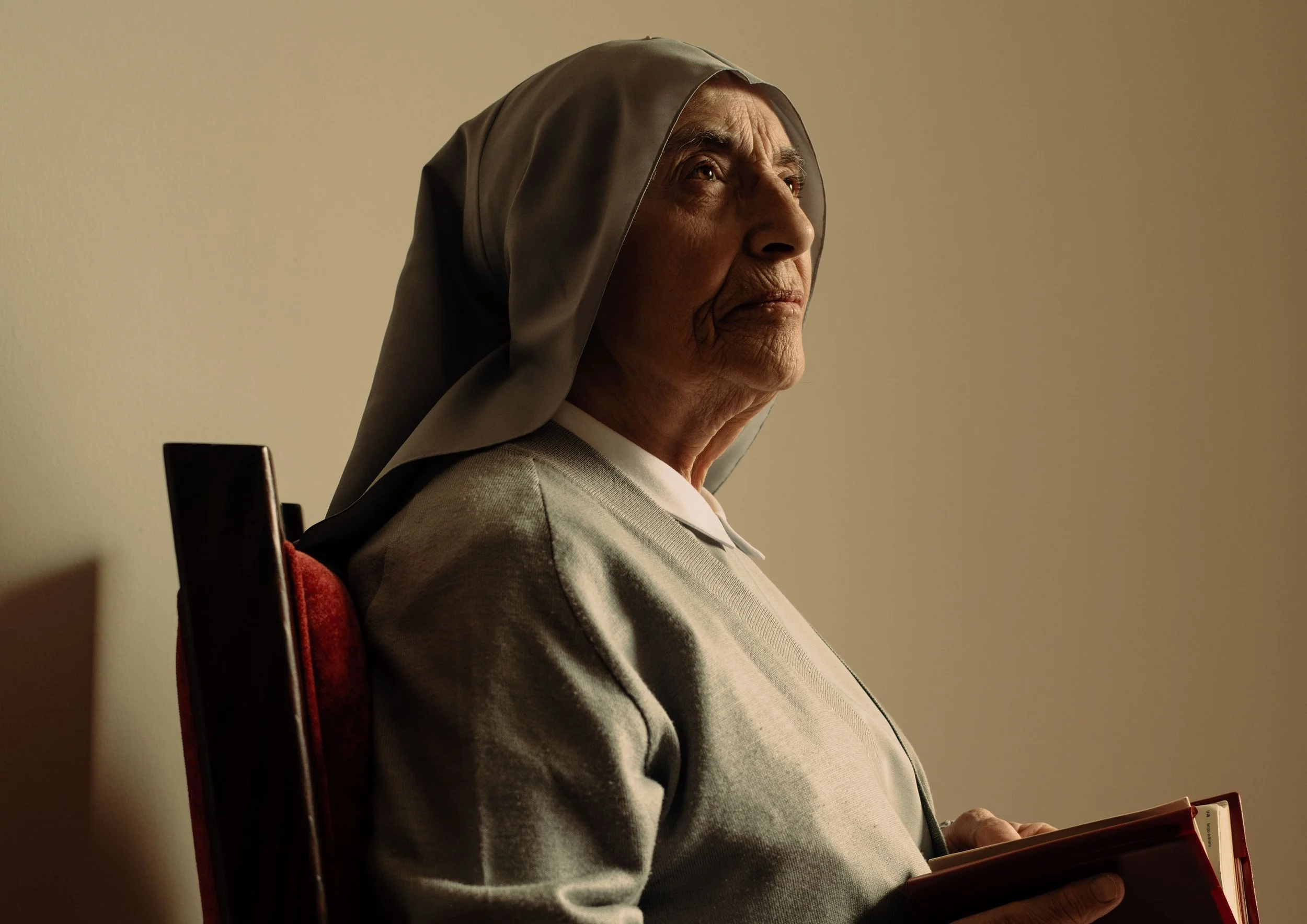 Elderly woman dressed as a nun, sitting in profile on a red chair, looking up thoughtfully while holding a book.