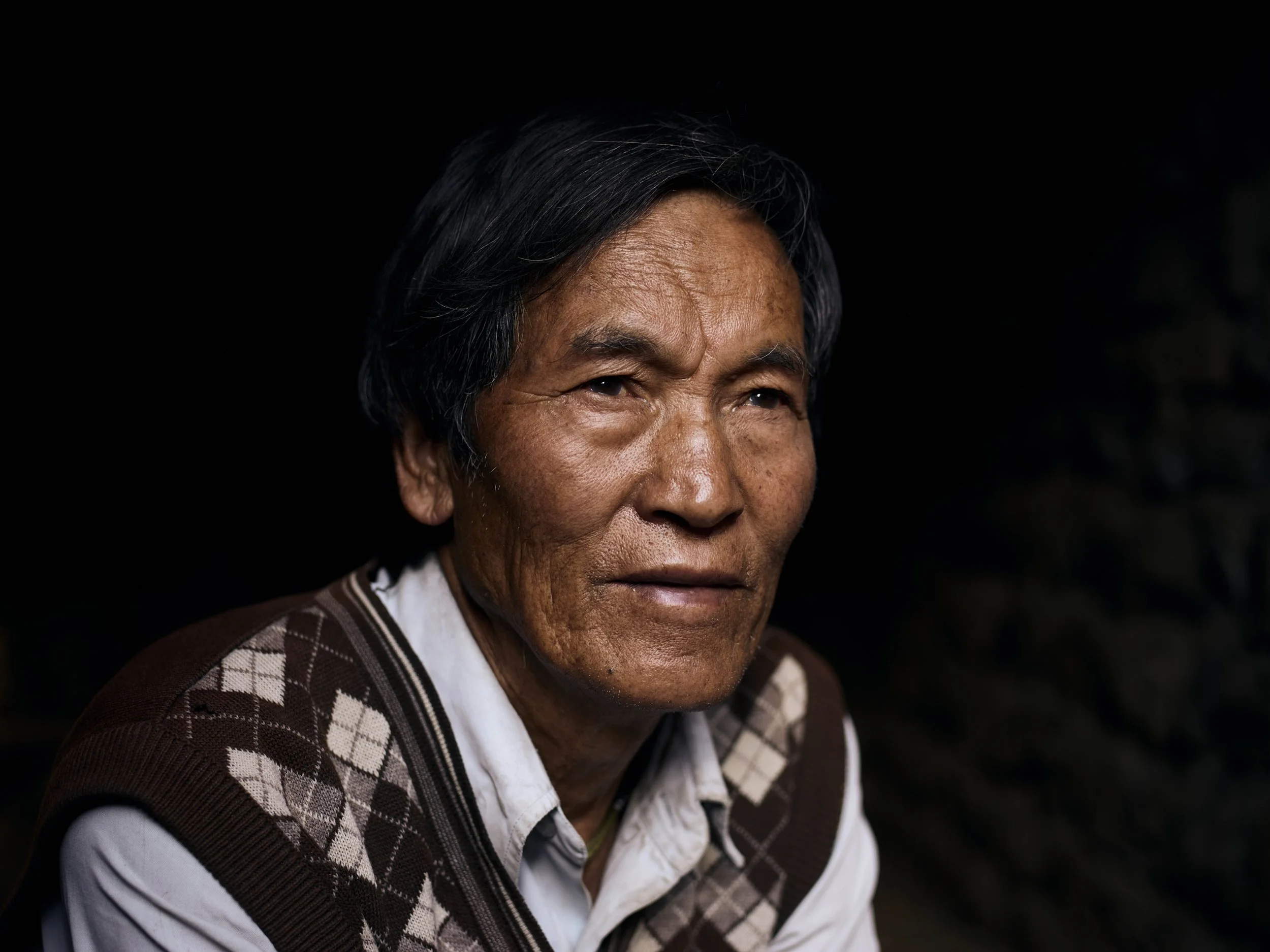 Close-up portrait of an elderly Asian man with black and gray hair, wearing a white shirt and a patterned brown vest, against a dark background.