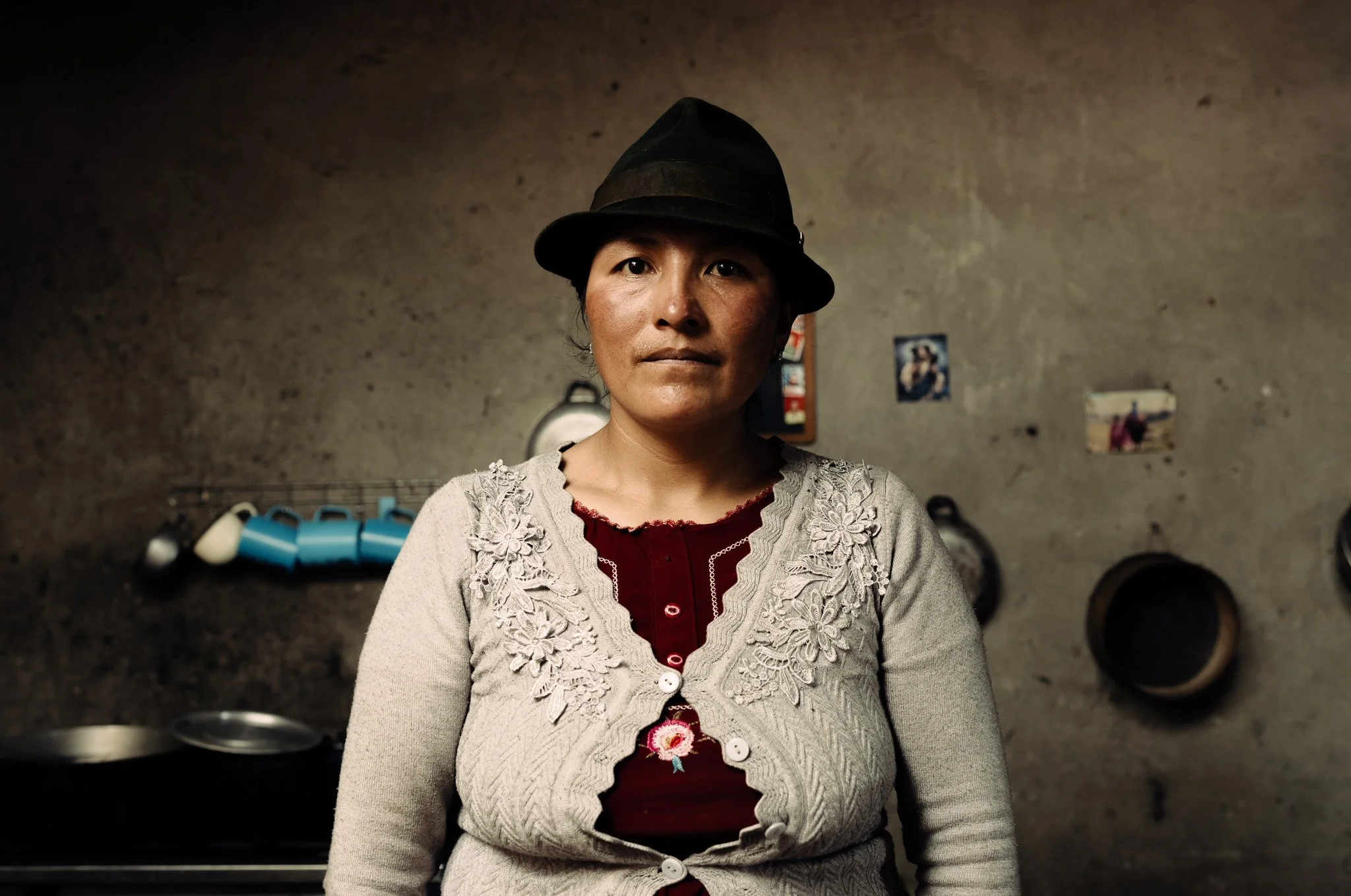 A woman wearing a dark hat and a beige embroidered cardigan standing in a rustic kitchen with a concrete wall, pots, and photographs on the wall.