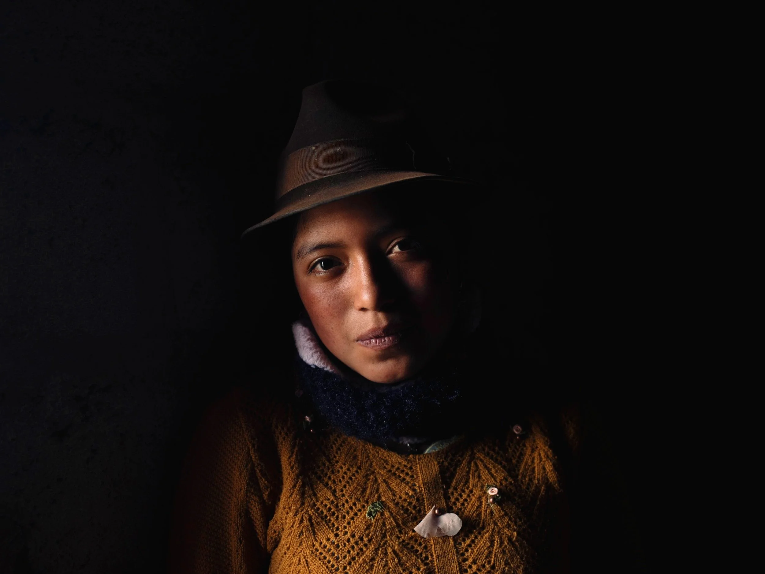 Portrait of a woman wearing a brown sweater, a dark scarf, and a hat, with dramatic lighting highlighting her face against a dark background.