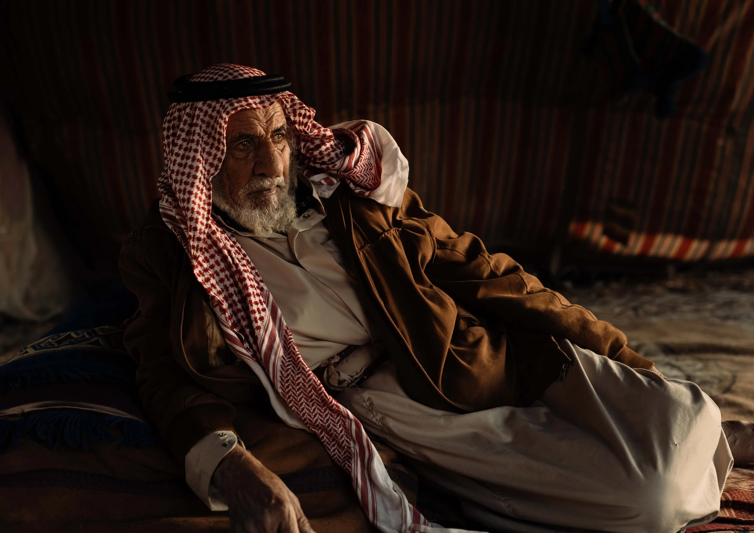 An elderly man with a white beard, wearing traditional Middle Eastern attire, including a red and white keffiyeh and brown cloak, is lying on a traditional woven bed inside a tent with striped fabric walls.