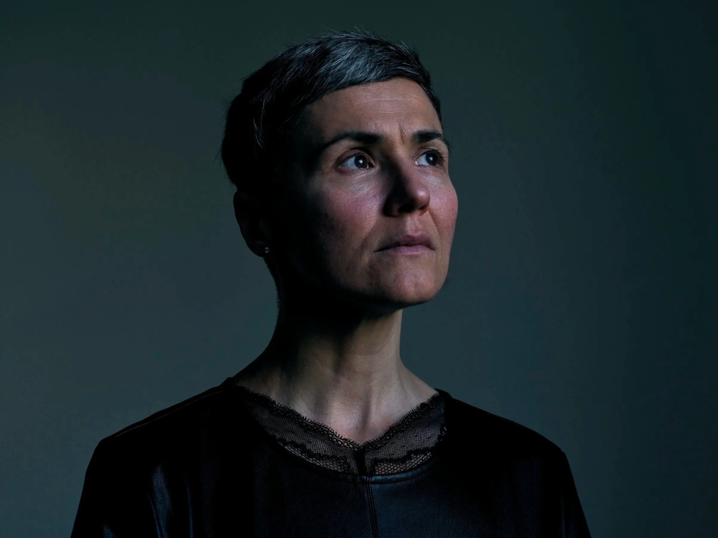 Portrait of a woman with short gray hair, looking slightly upward. She has a serious expression and is wearing a black top with a lace neckline. The background is dark and plain.