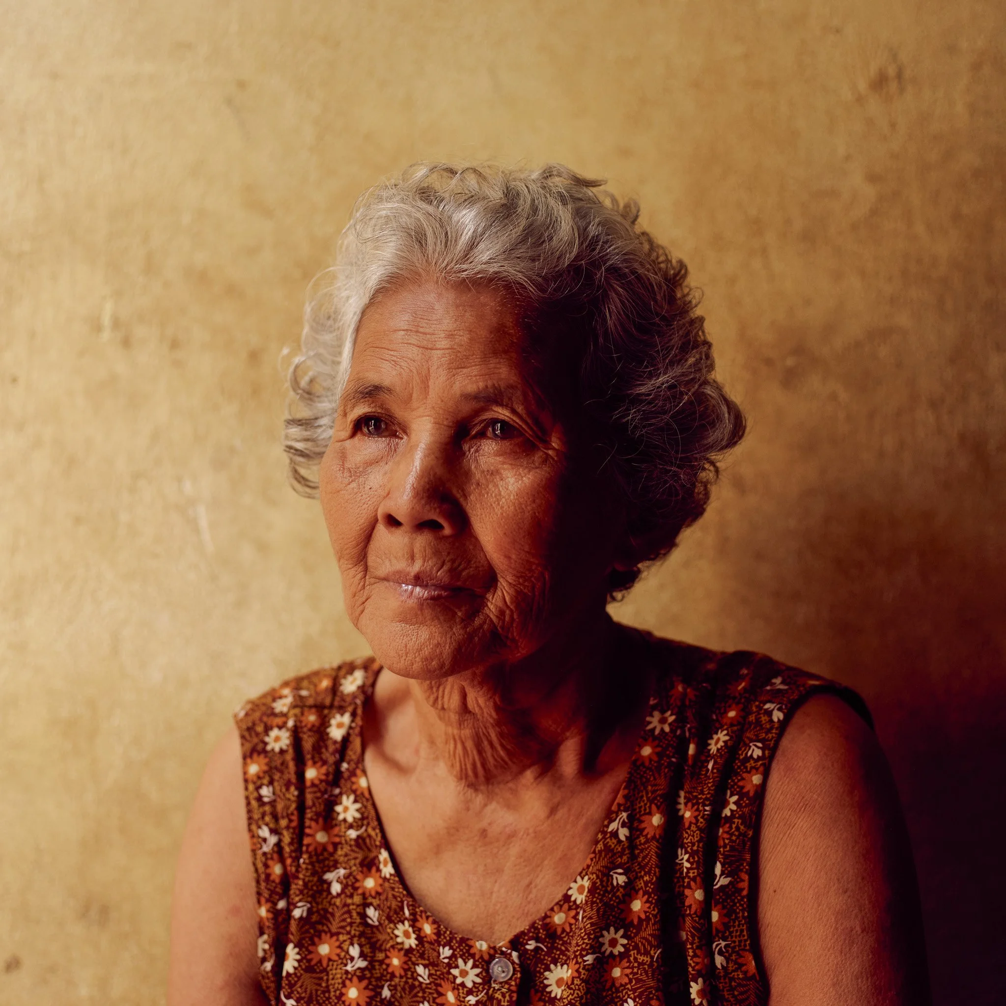 An elderly woman with short, curly, gray hair looks thoughtfully to the side, wearing a brown floral sleeveless top against a plain beige background.
