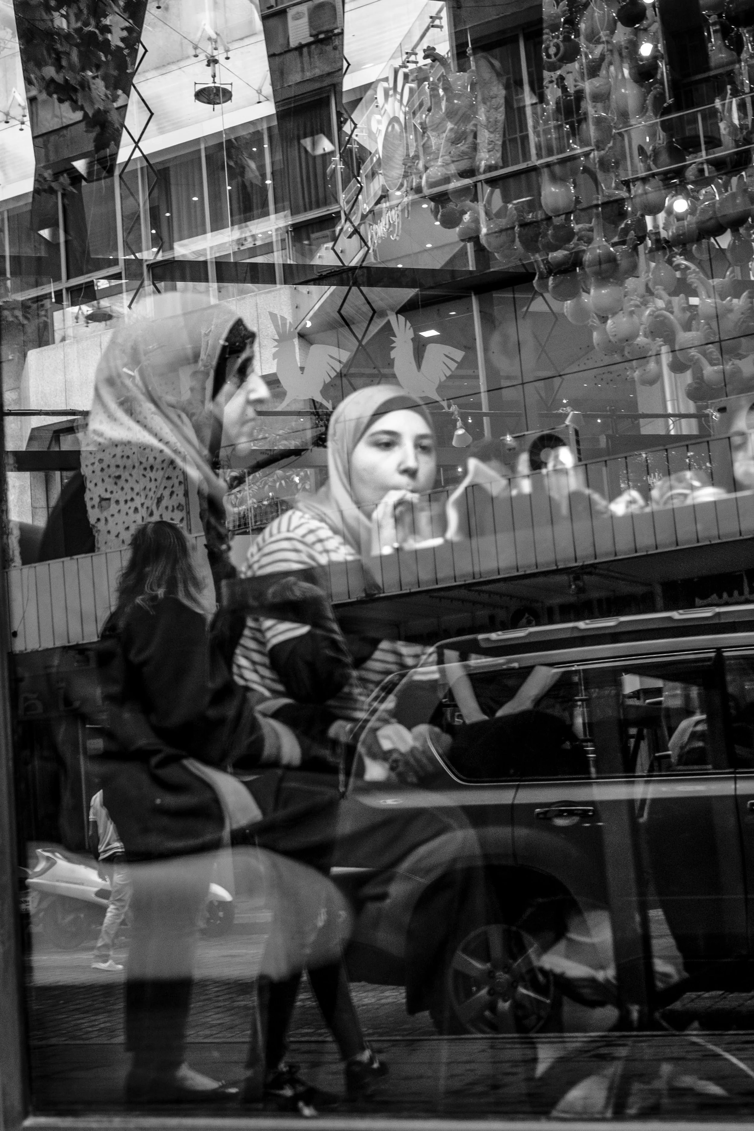 Reflejo de una escena urbana en un cristal con varias personas caminando, incluyendo dos mujeres con hijas pequeñas y un auto en la calle.