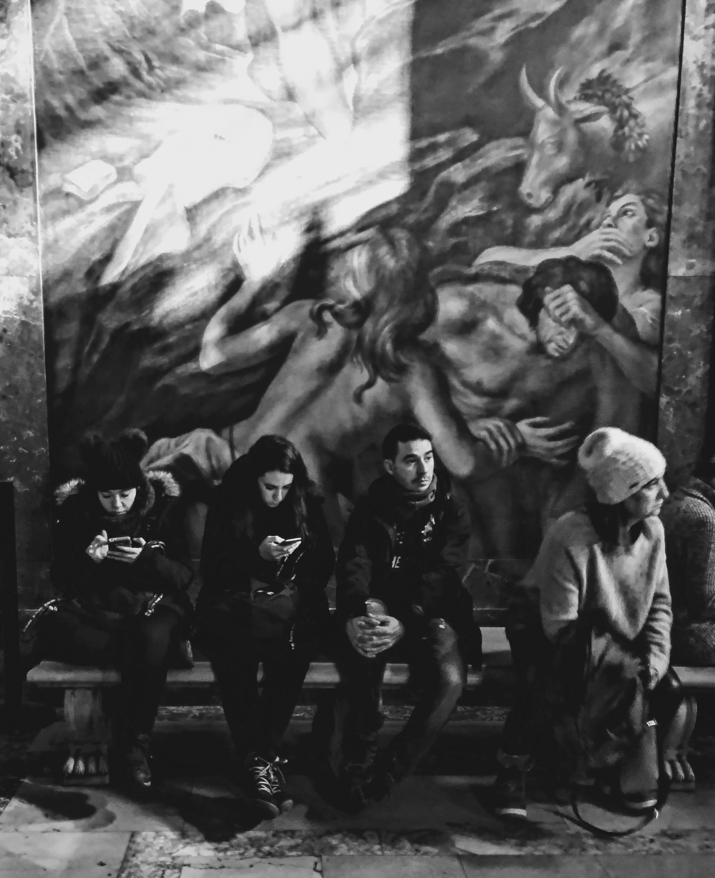 Un mural en blanco y negro de un caballo con un cuerno en su cabeza y una figura humana con el brazo extendido, con varias personas jóvenes en primer plano usando ropa oscura, algunas con teléfonos móviles en las manos.