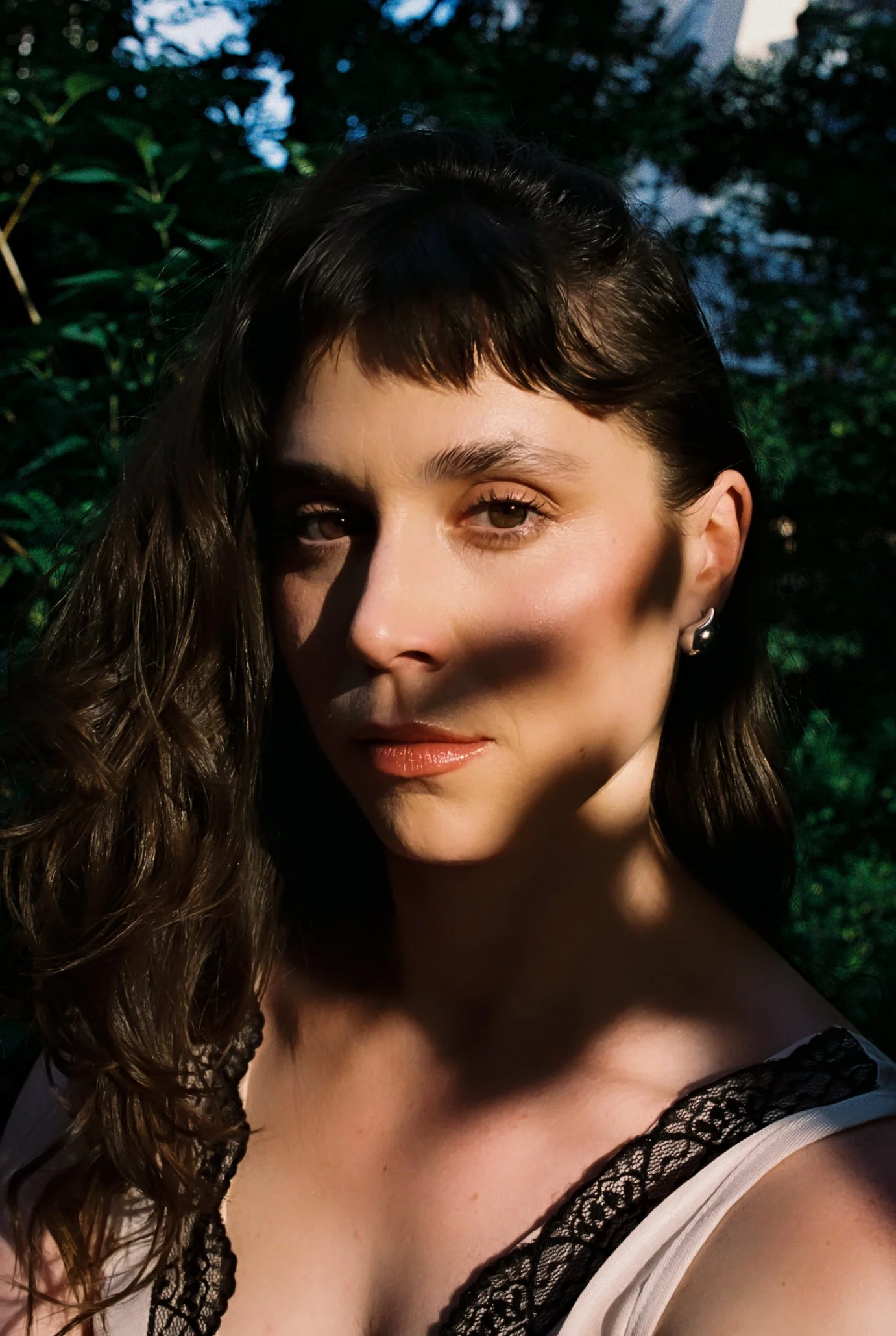 Retrato de una mujer con cabello rizado, de piel clara, en un entorno natural con árboles y cielo al atardecer.