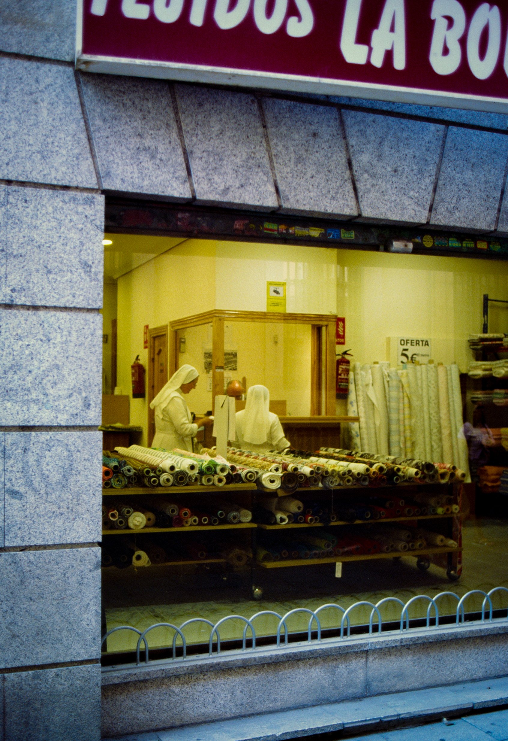 Vista de una tienda de cortinas con varias telas enrolladas en estantes de madera y una empleada atendiendo a un cliente desde el interior, visible a través de la ventana.