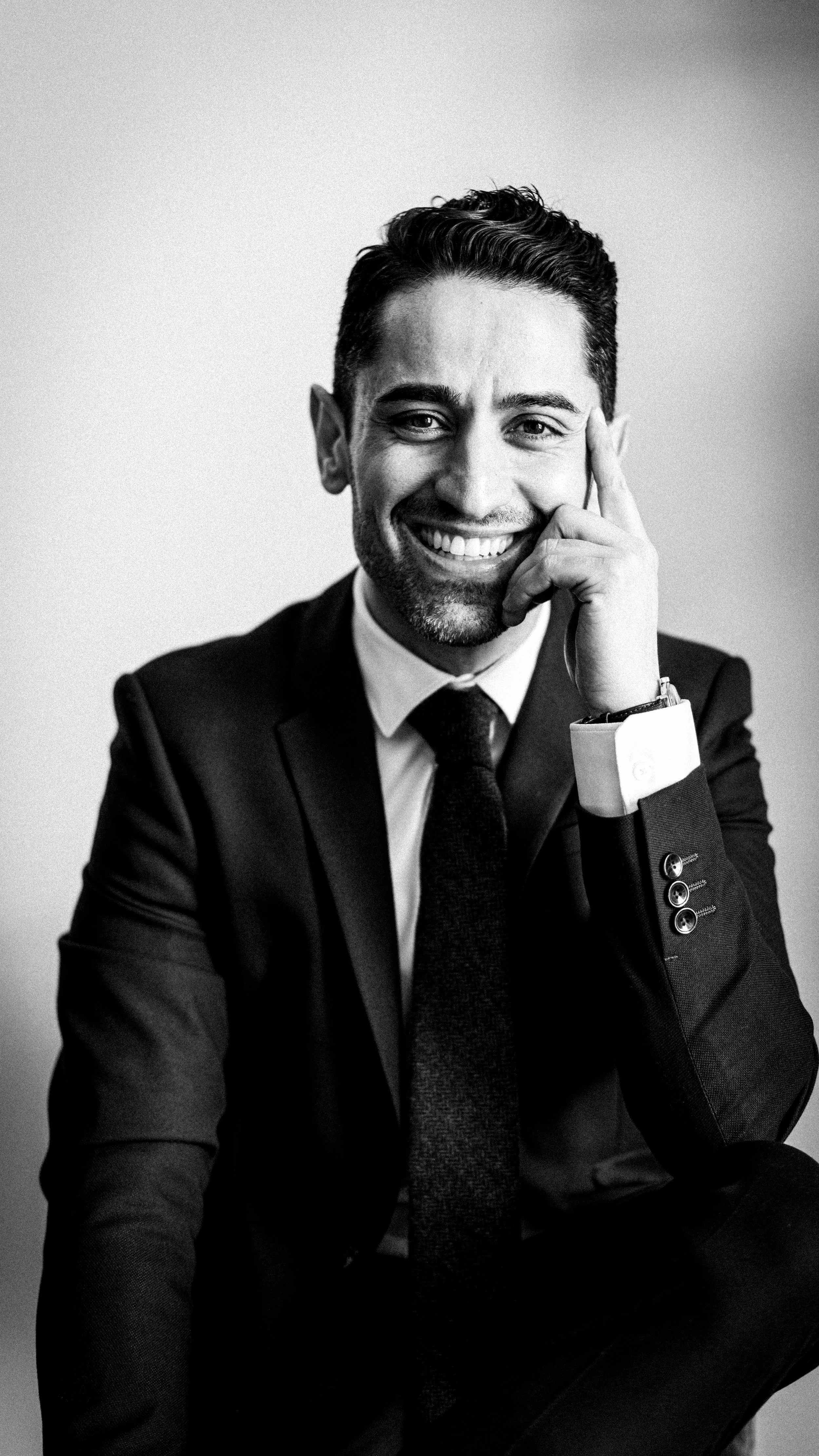 Retrato en blanco y negro de un hombre sonriendo, vestido con traje y corbata, sentado con la mano en la cara y usando un reloj