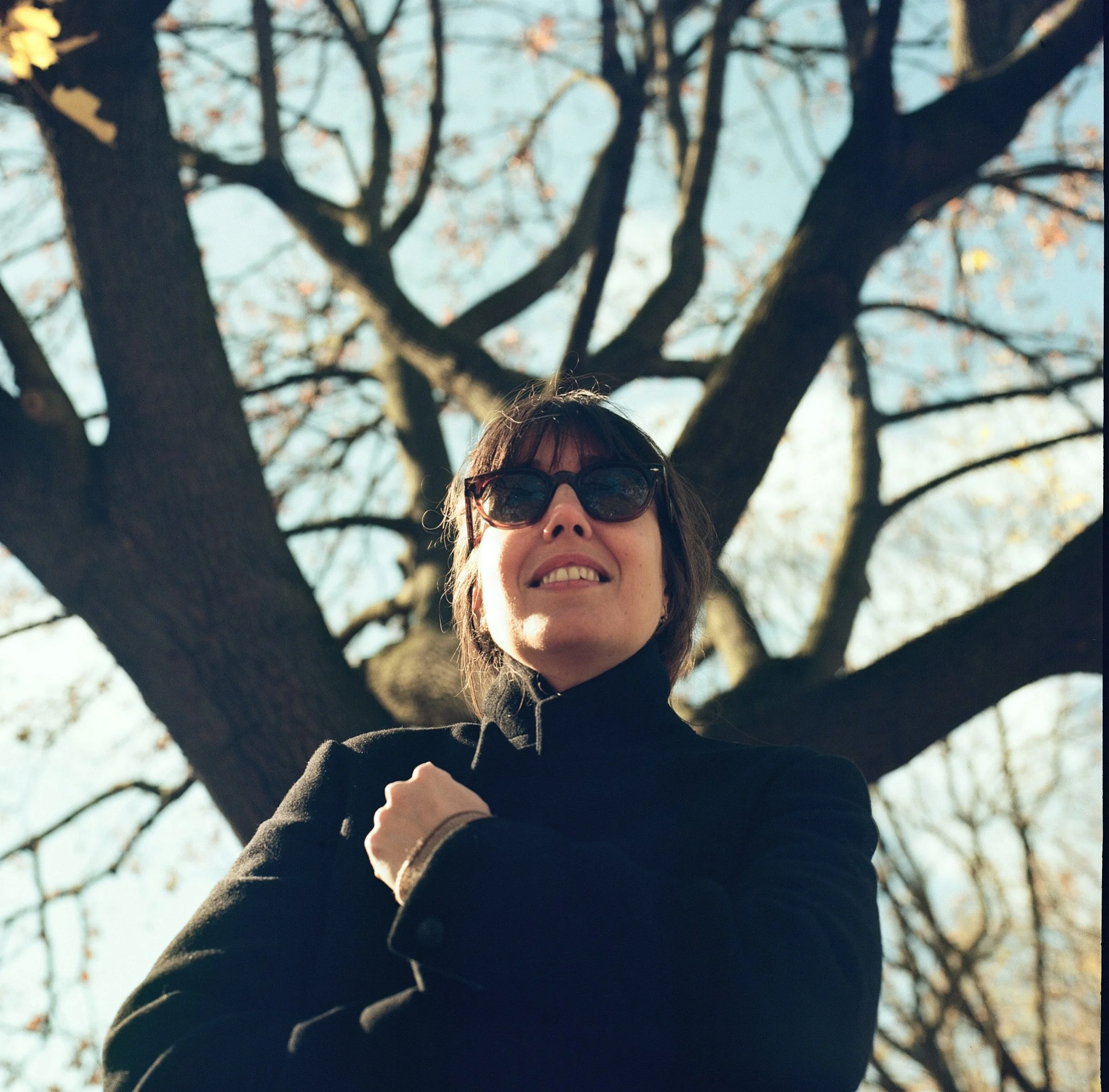 Una mujer con gafas de sol de cabello castaño, usando un suéter negro, está de espaldas en un árbol de octubre o noviembre, con ramas desnudas y algunas hojas amarillas.