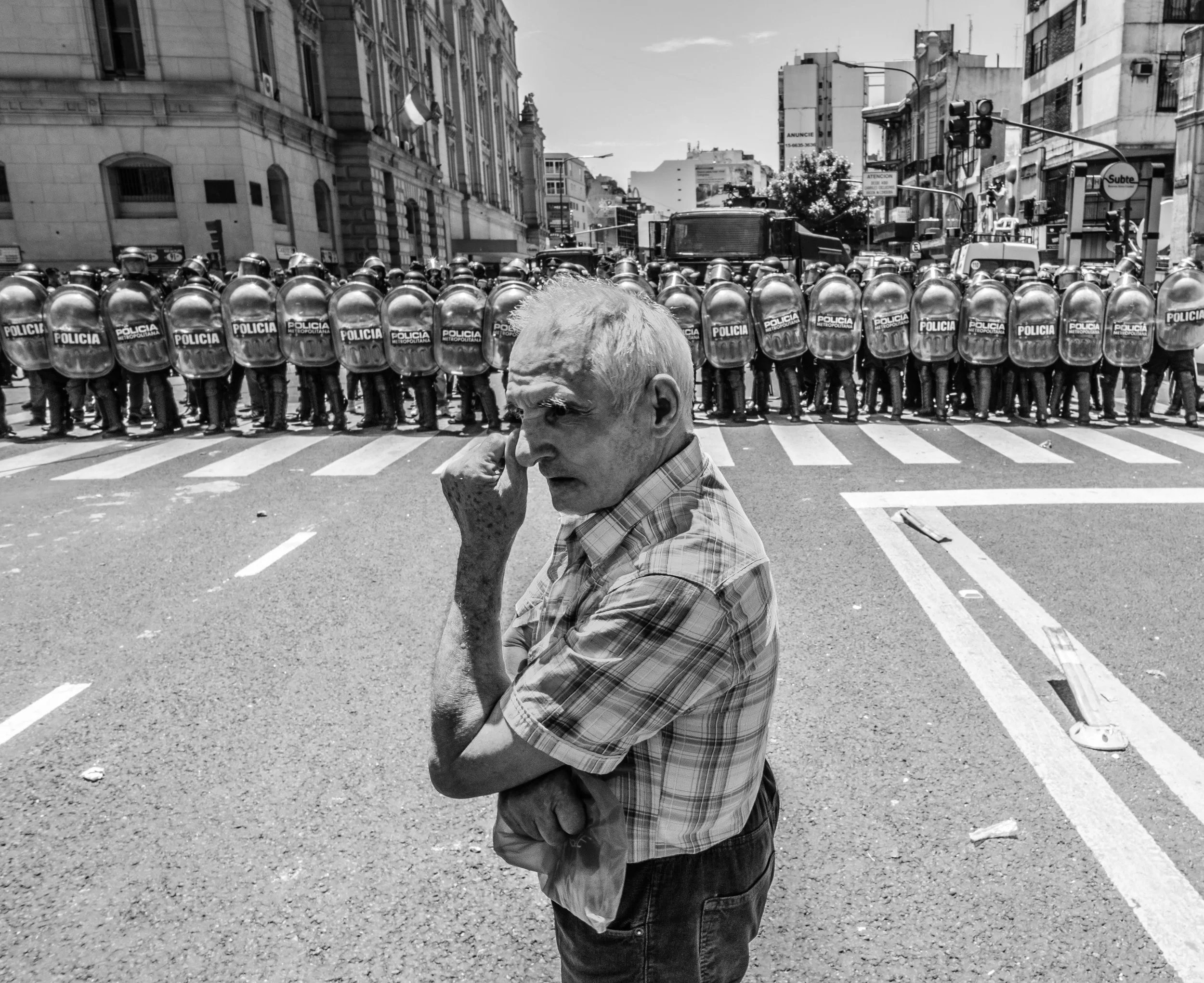 Un hombre mayor con cabello canoso, vestimenta a cuadros, sostiene su mejilla con una mano mientras camina por una calle. En el fondo, una línea de policías con cascos y escudos formando un bloqueo en la calle de una ciudad con edificios modernos y c