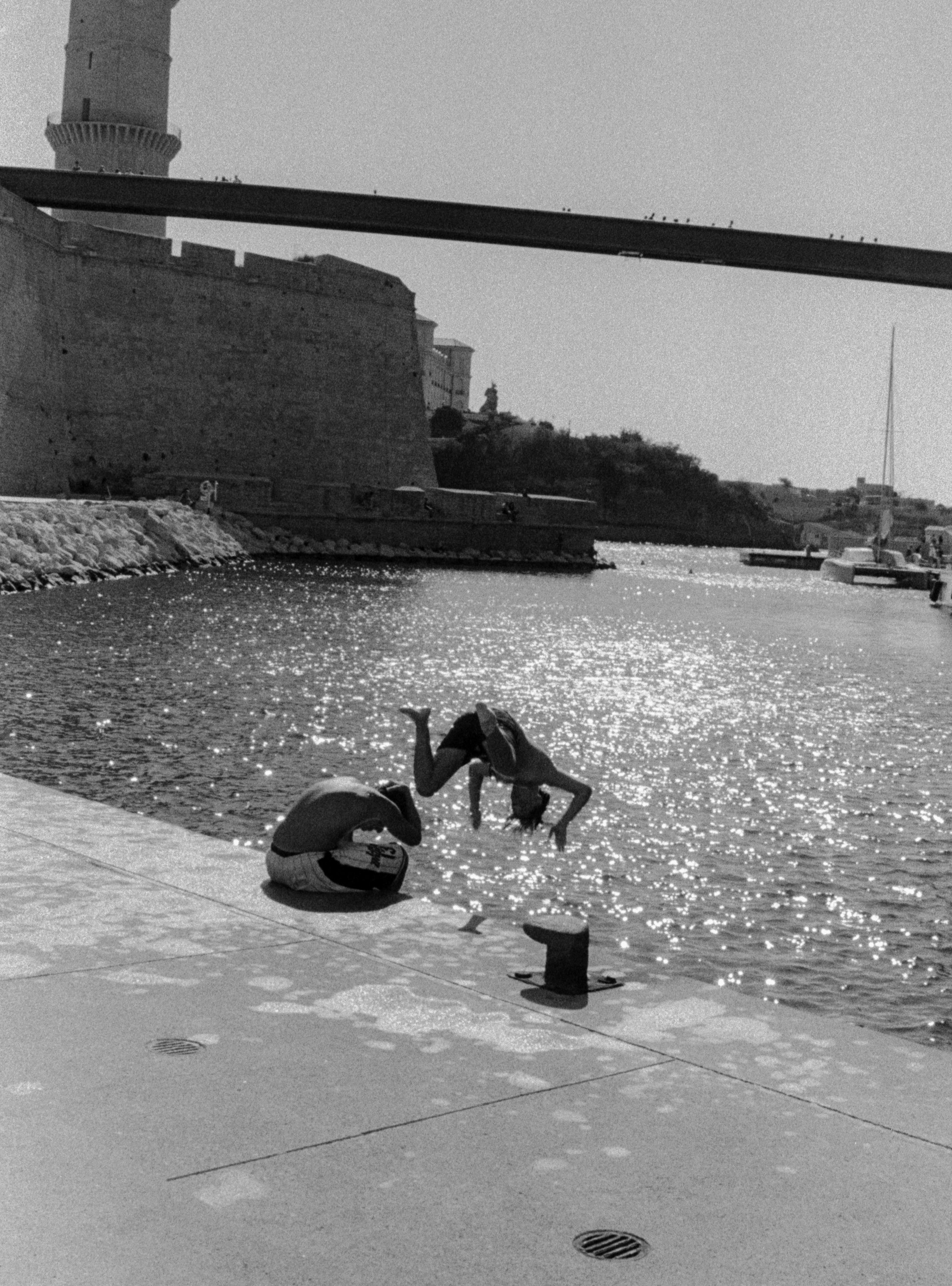 Una persona agachada por el agua en un muelle junto a un río, con un monumento y estructuras con vista al agua, en una imagen en blanco y negro.