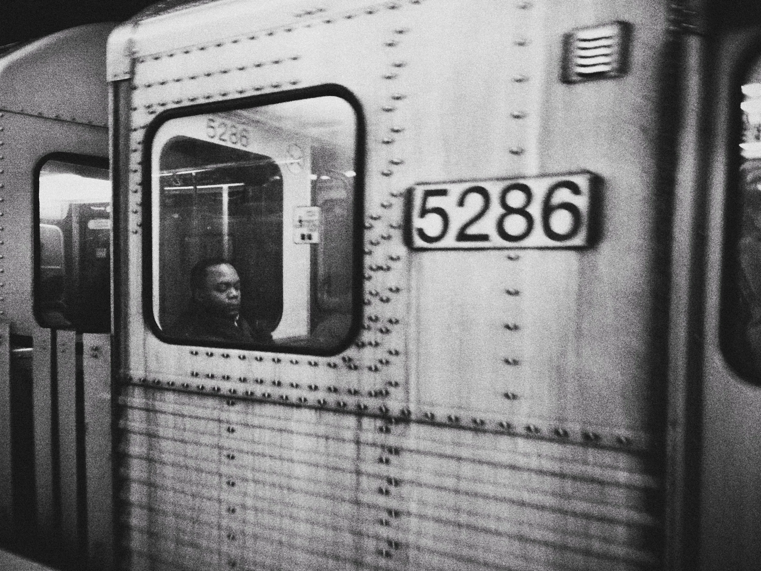 Un camión de carga grande con matrícula 5206. En la esquina izquierda se ve una persona observando por la ventana del camión. La imagen es en blanco y negro y tiene un aspecto borroso.