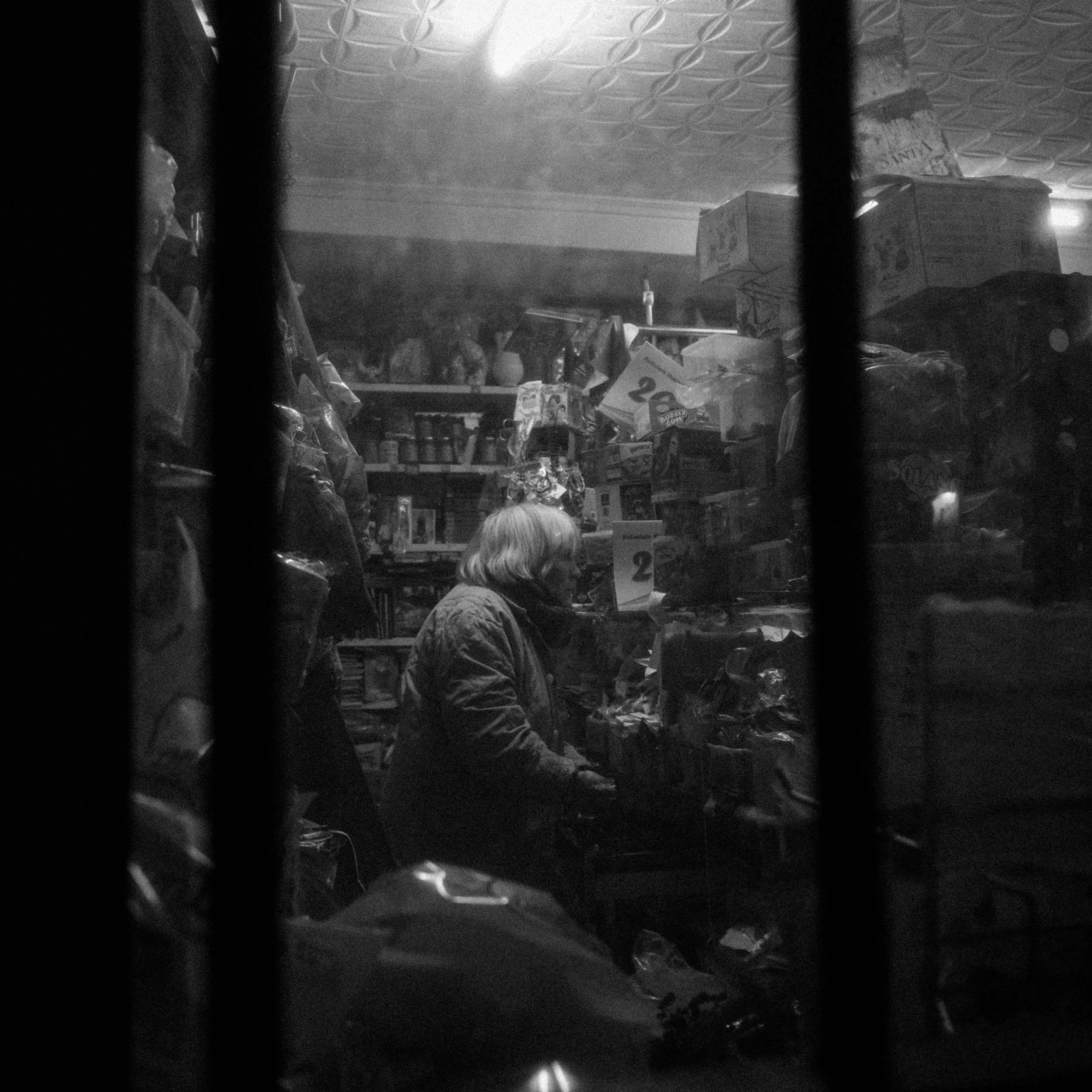 Vista desde afuera de una tienda con productos en estantes, vista a través de barrotes o reja, en la tienda hay cajas y paquetes de comida y otros artículos.