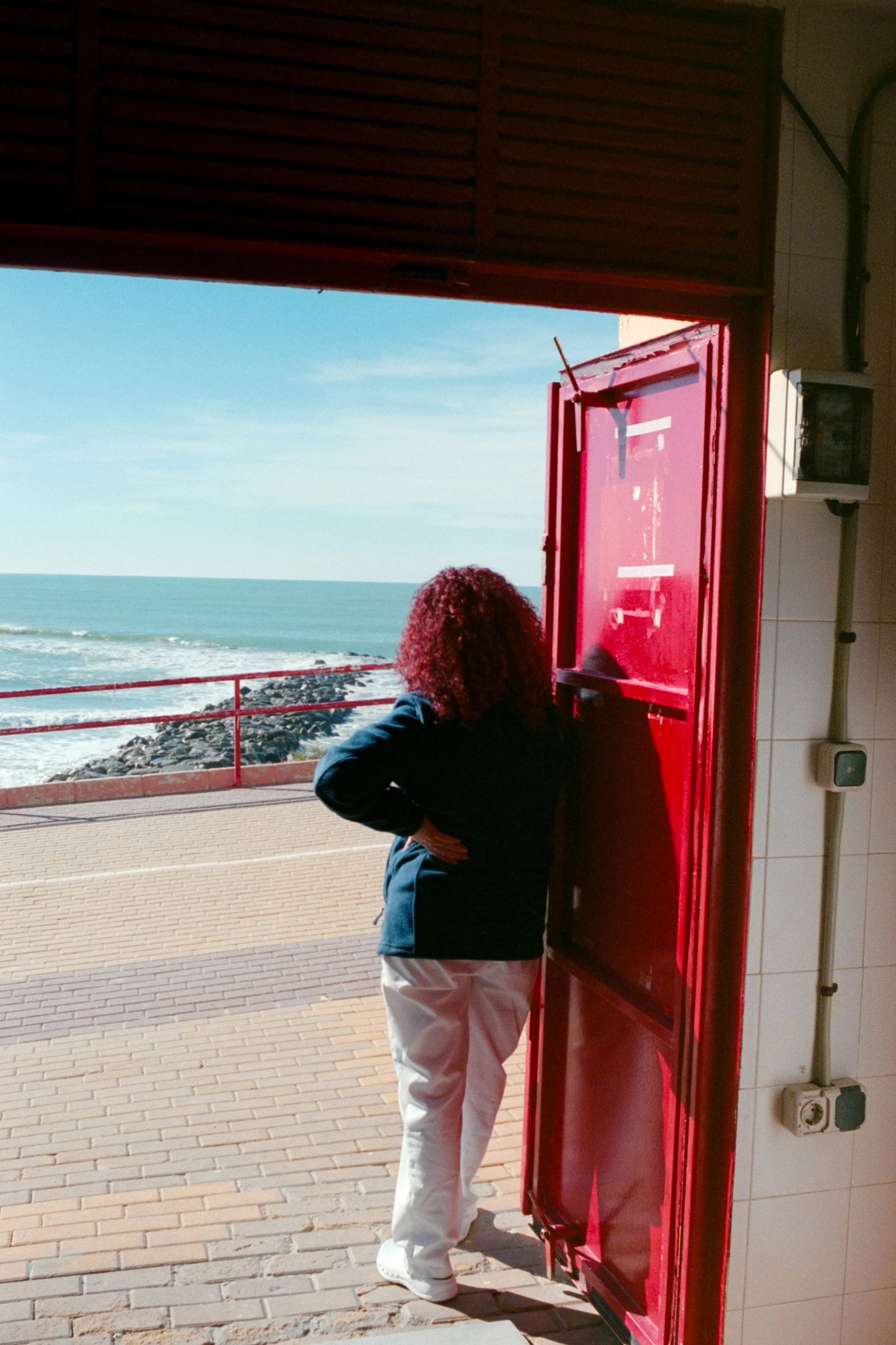 Persona con cabello rizado y ropa de color oscuro, de espaldas, frente a una puerta roja en un área junto a la playa y el mar. La puerta está abierta y hay un espacio en la pared con enchufes y un interruptor.