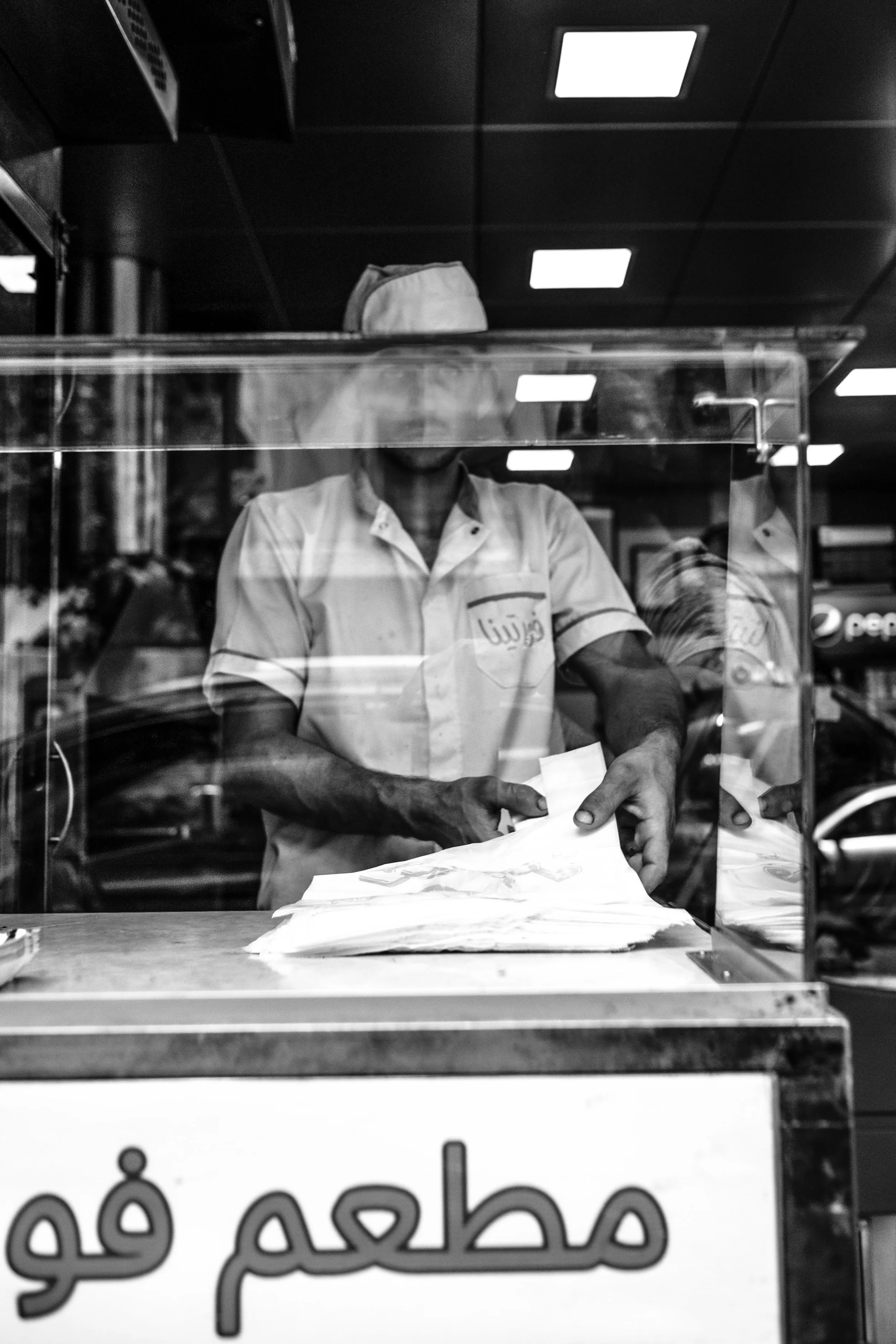 Hombre sirviendo comida en un restaurante de comida rápida desde detrás de un mostrador de vidrio, con letrero en árabe en la parte inferior de la imagen.
