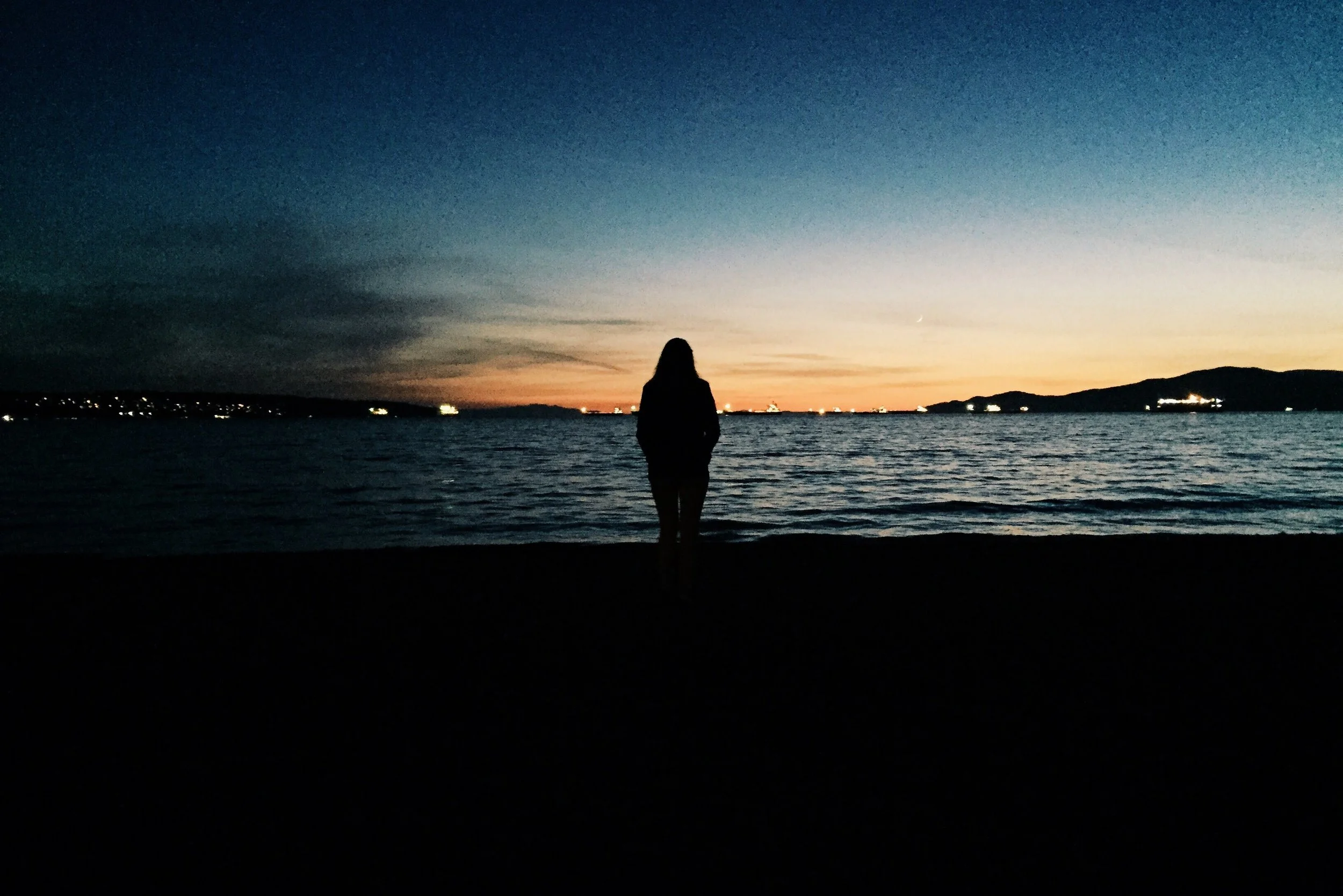 Persona de espaldas observando el mar durante el atardecer, con cielo oscuro y luces en la costa.