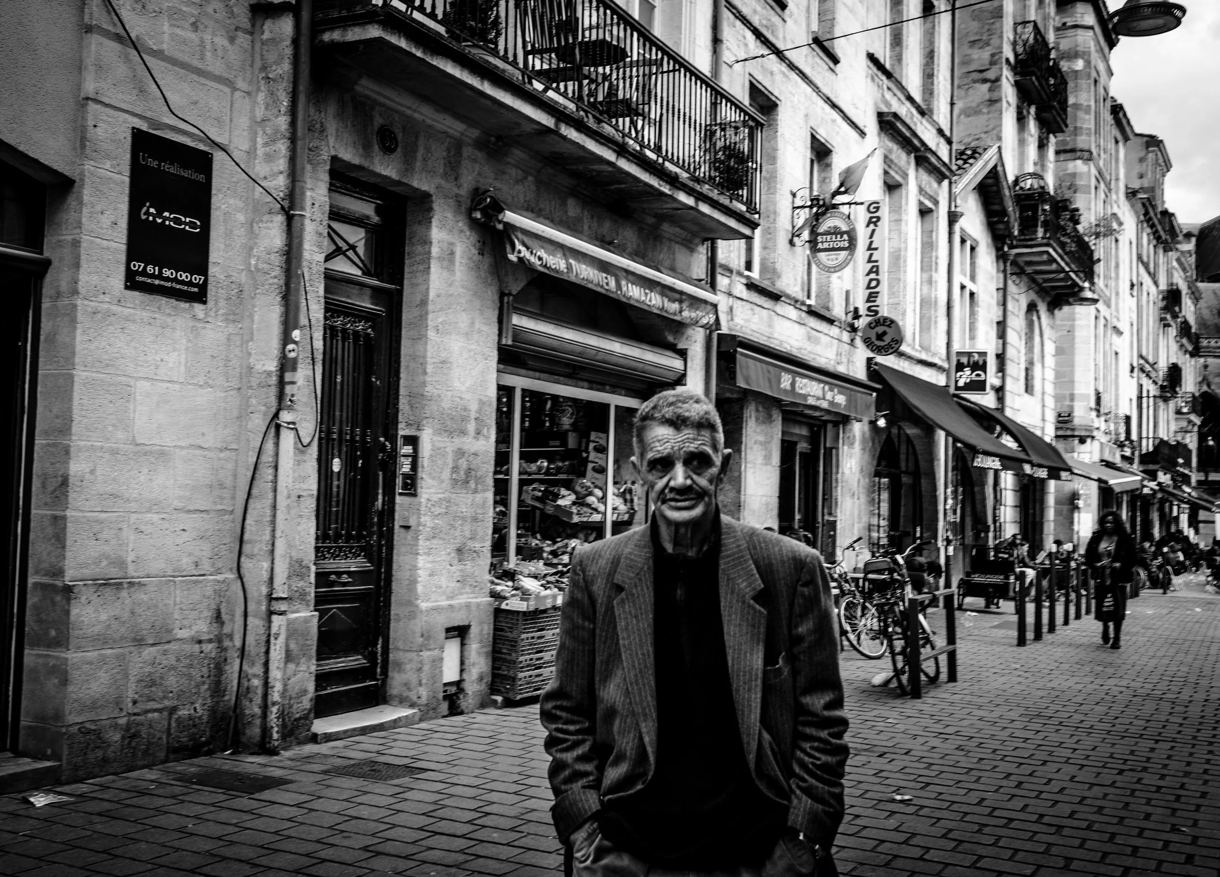 Hombre con traje gris caminando por una calle peatonal en una ciudad. Edificios antiguos, balcones y señalización visible. Ambiente urbano en una ciudad europea.