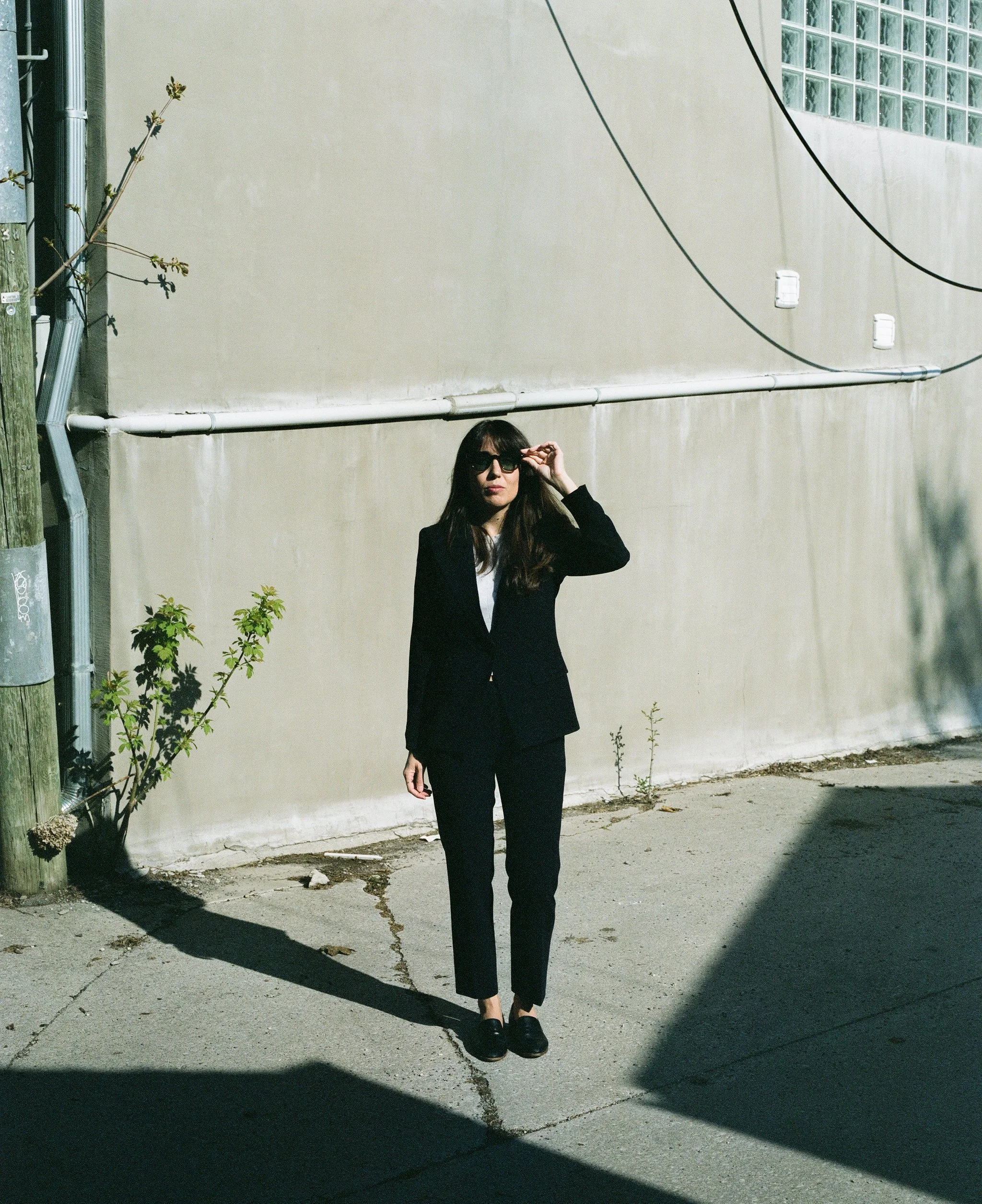 Una mujer con gafas negras, ropa negra y zapatos negros, de cabello largo y oscuro, posa contra una pared beige en la calle, bajo cables eléctricos y sombra en el suelo.