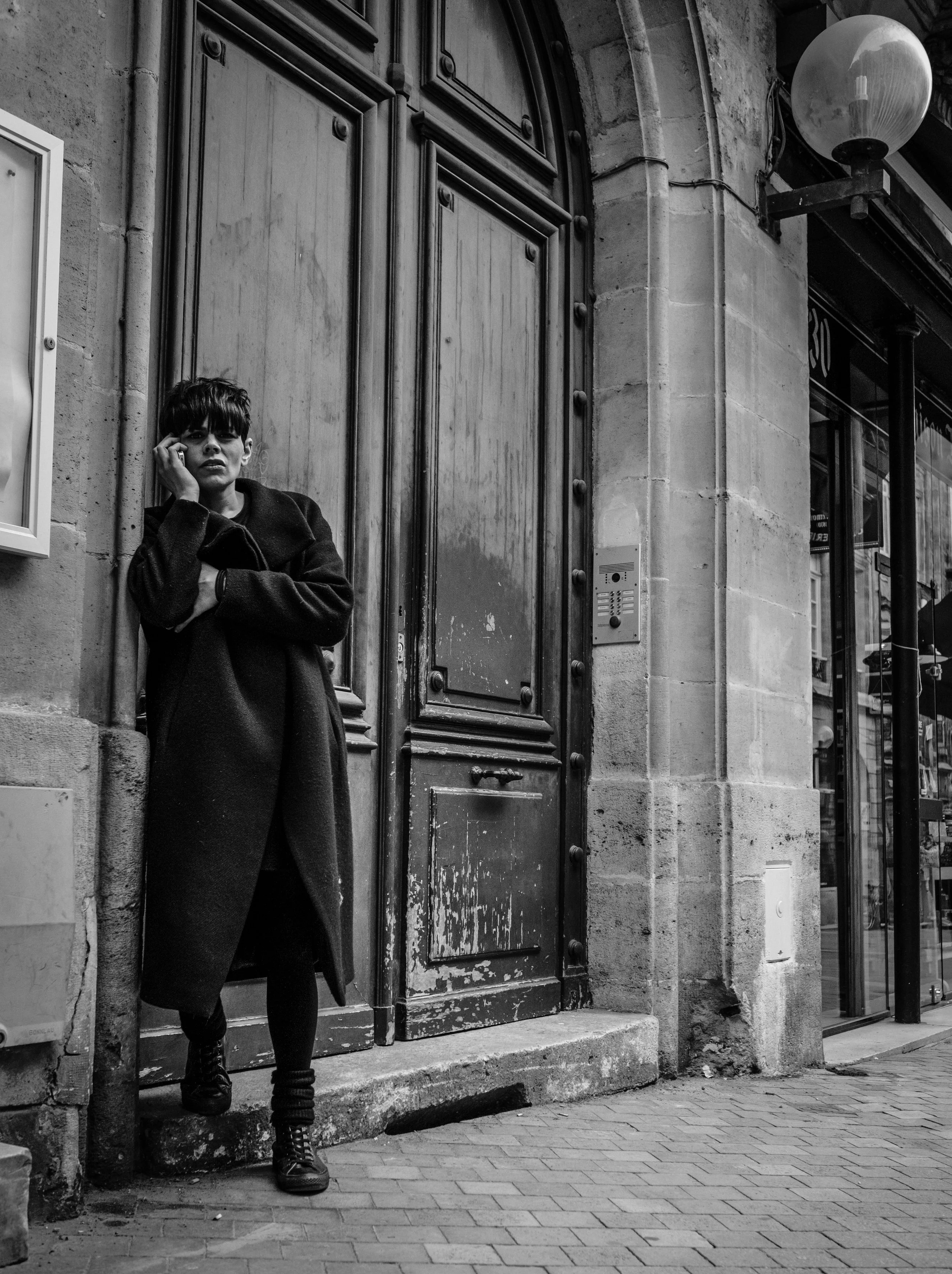 Joven de pie junto a una puerta de madera en la calle, llevando una chaqueta oscura y botas, con el cabello oscuro y corto, en un entorno urbano en blanco y negro.