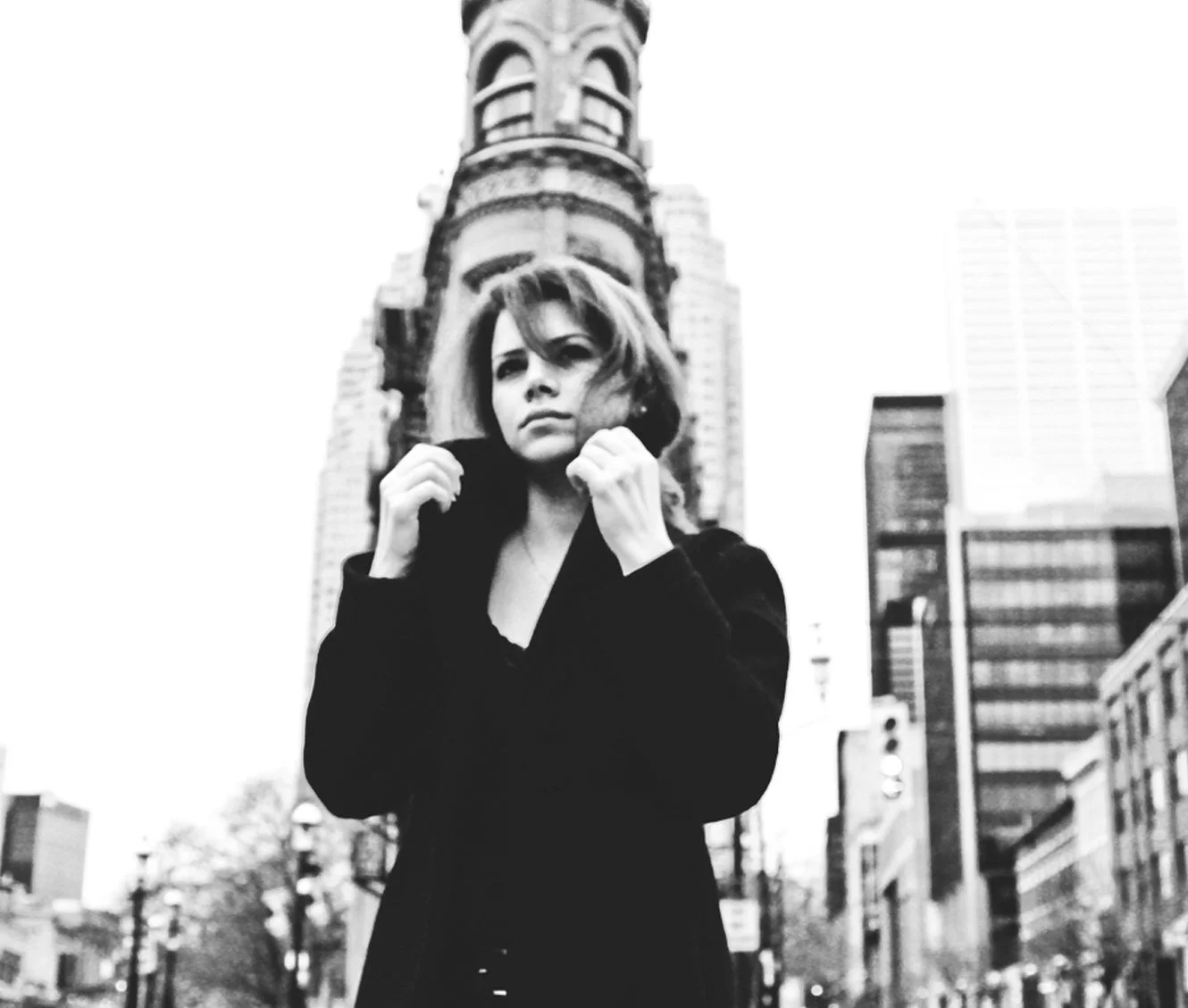 Mujer con ropa negra al frente de edificios en ciudad, en blanco y negro.