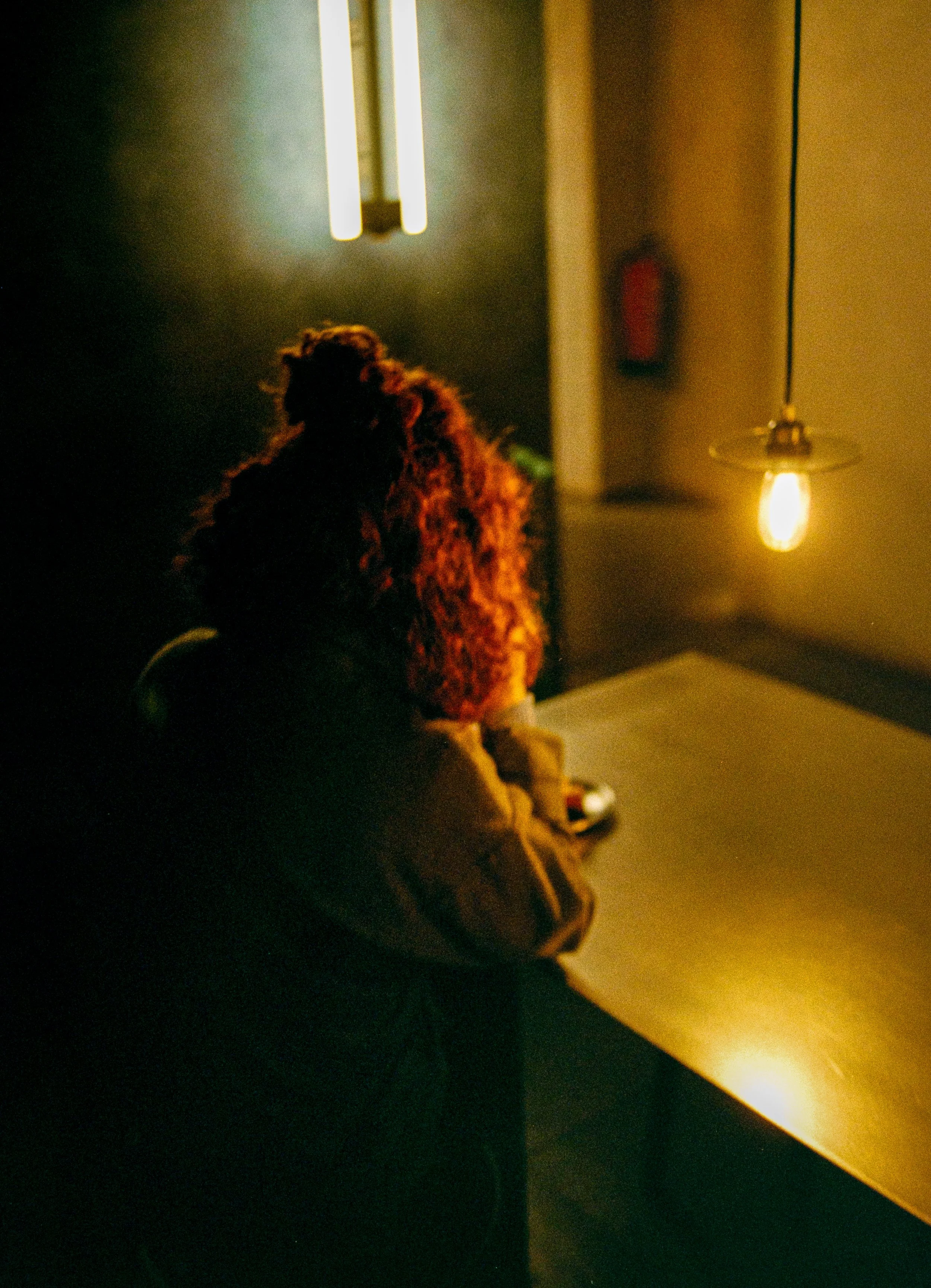 Una mujer con cabello rizado oscuro sentado en una mesa, en un ambiente con iluminación tenue, una lámpara colgante y un fondo con una pared oscura y una luz brillante.