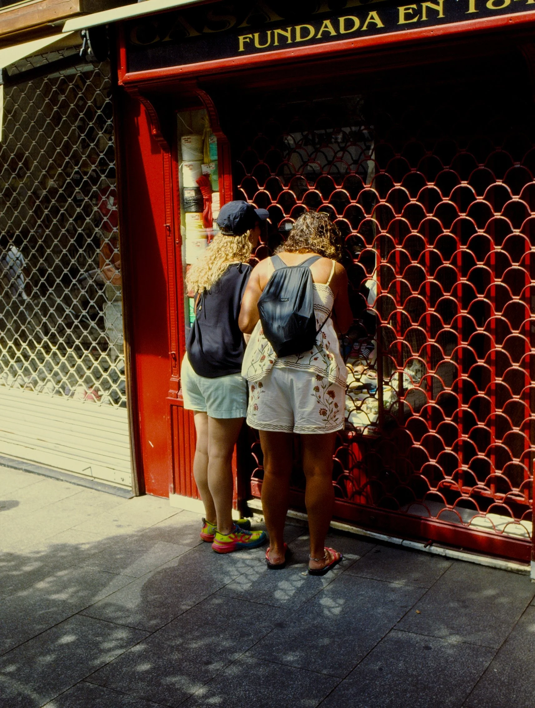 Dos mujeres con mochilas observando un escaparate con rejas rojas en una calle. La tienda está cerrada y en el cartel se lee 'Fundada en T'.