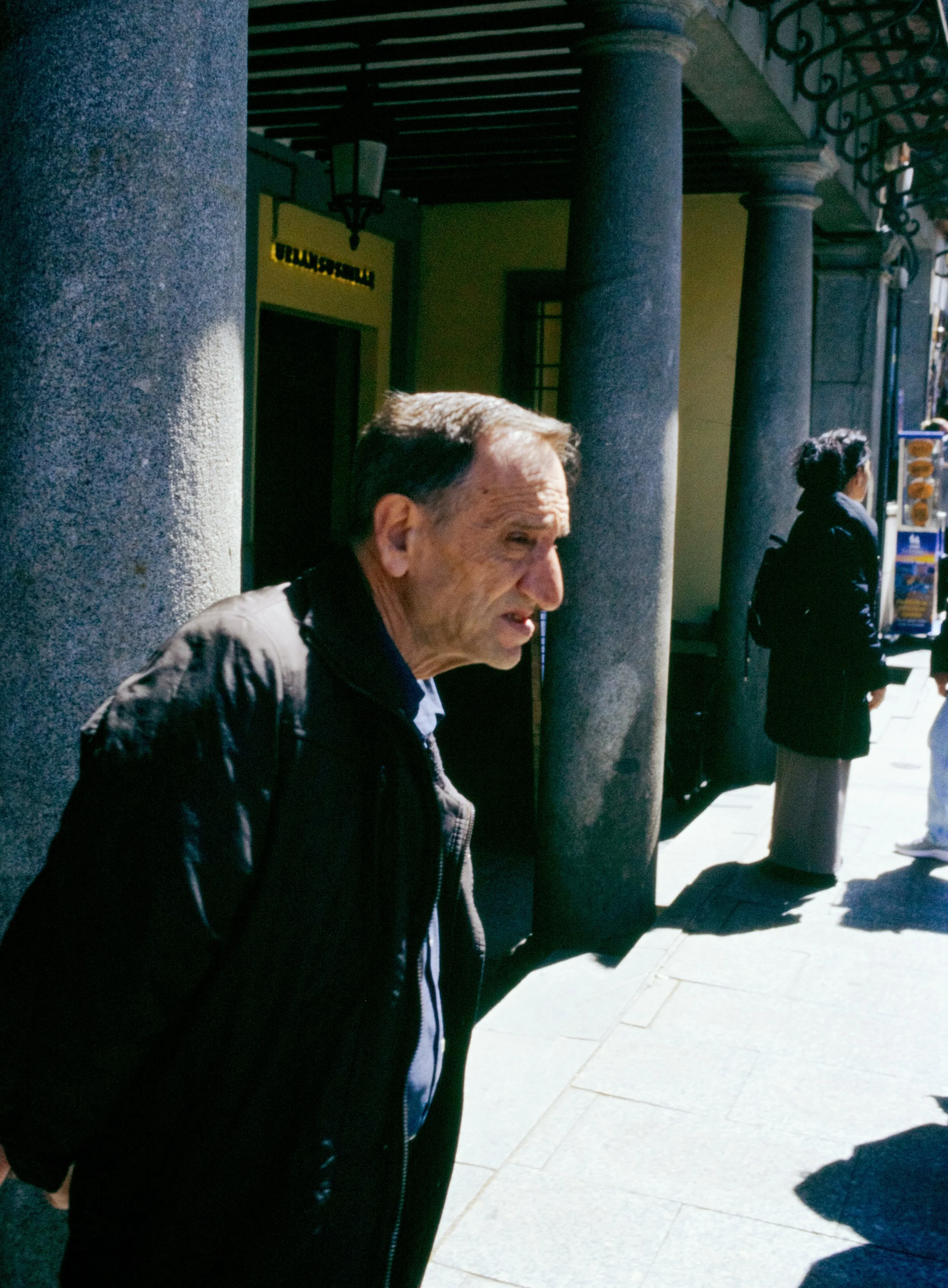 Un hombre y una mujer de espaldas y a la mitad del cuerpo, en una calle, con columnas y luz solar, parecen estar esperando o conversando.