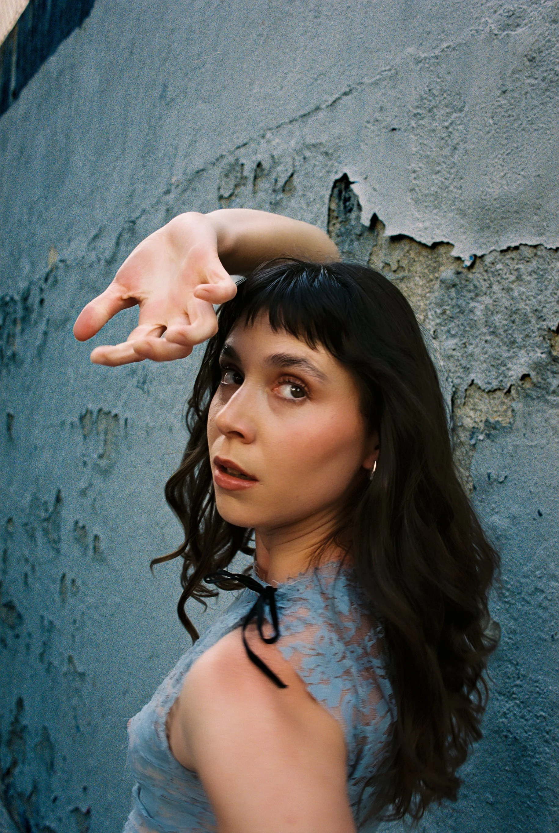 Mujer de cabello castaño oscuro con ojos marrones, posando junto a una pared de concreto gris, vestida con blusa de encaje azul y movimiento en su cabello.