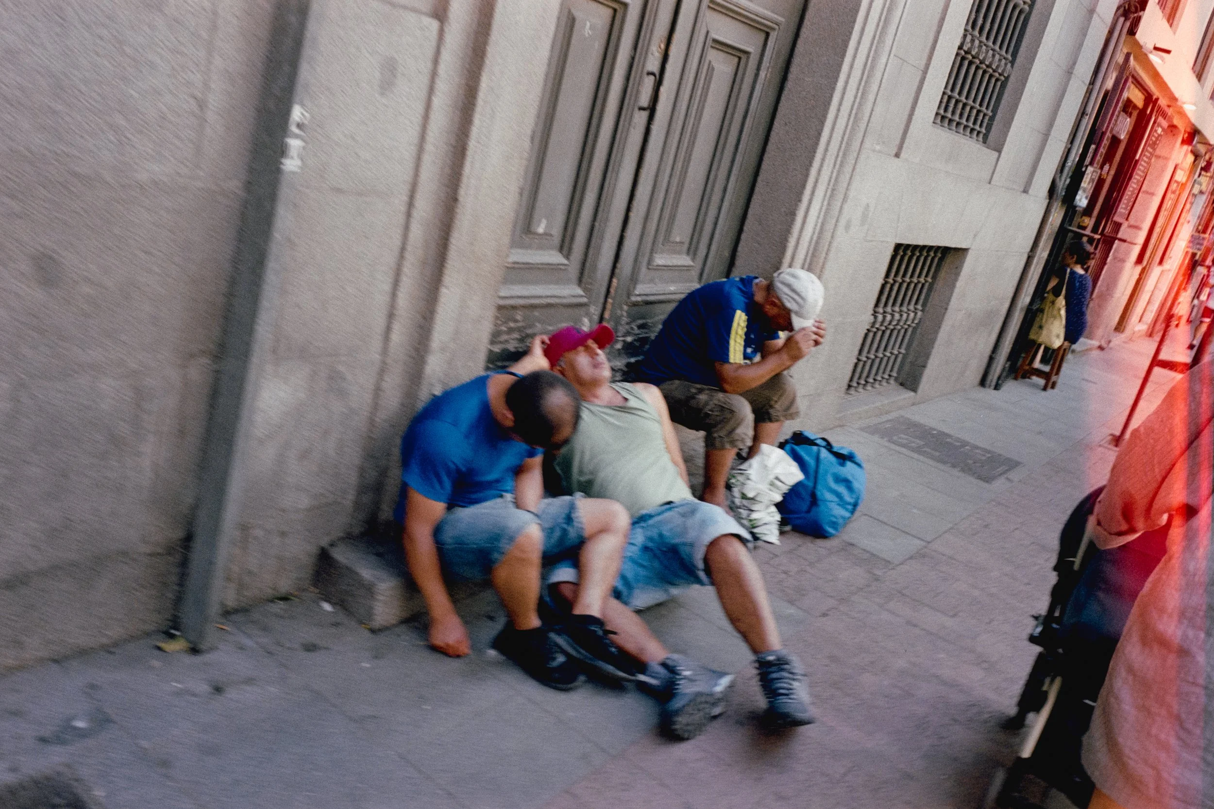 Varias personas descansando y durmiendo en la acera de una calle urbana, con algunos objetos personales a su lado, en un entorno urbano durante el día.