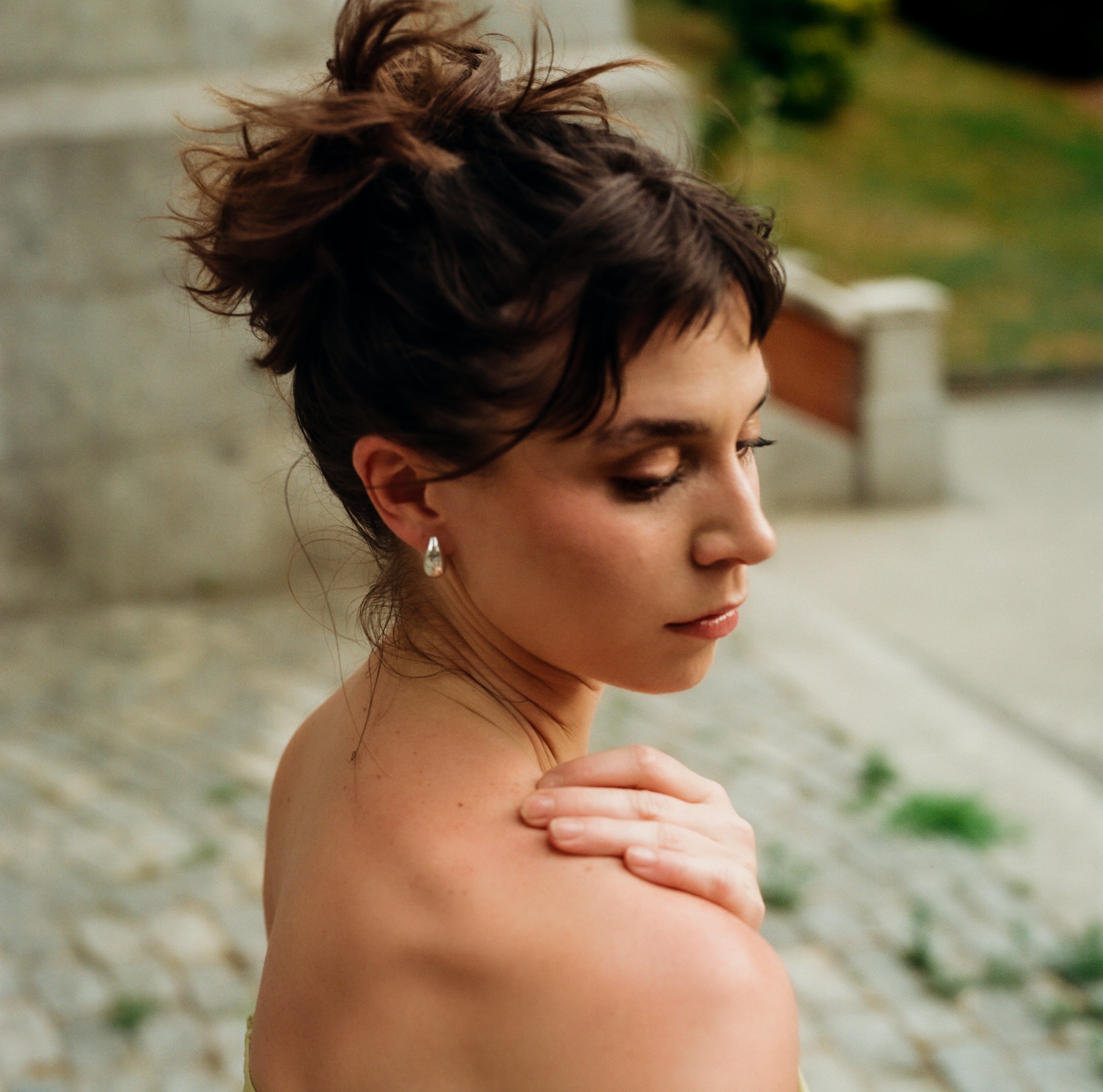 Mujer con cabello oscuro y ondulado, con un peinado recogido en la parte superior, cubriéndose suavemente con una mano sobre el pecho, en un entorno de fondo borroso con tonos verdes.