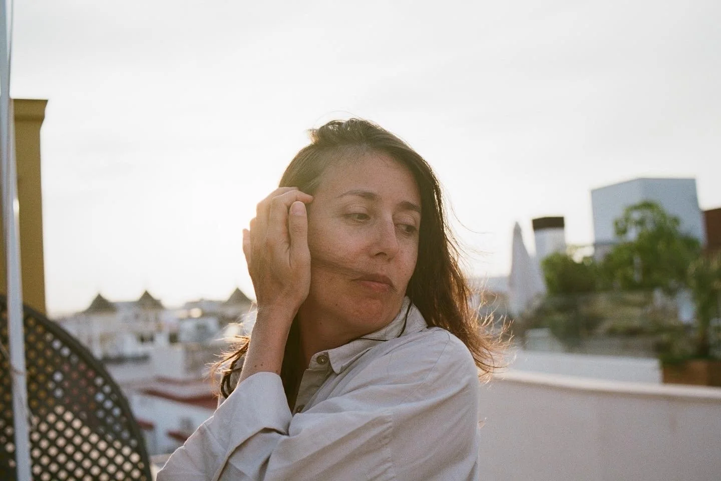 Mujer con cabello largo y suelto, vistiendo una camisa blanca, con vista a edificios y árboles al atardecer.