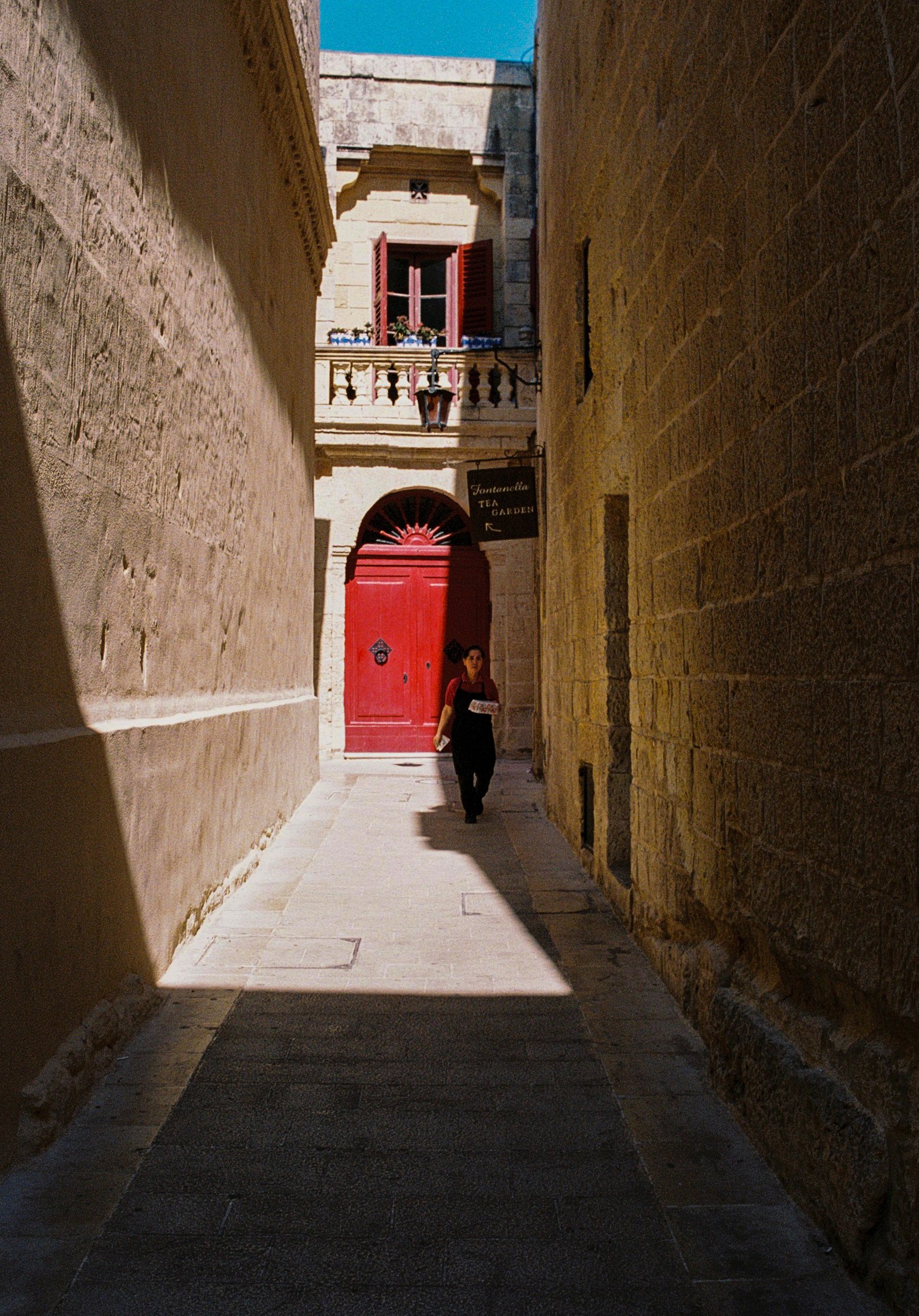 Un callejón estrecho con paredes de piedra, sombra en la parte inferior y luz que entra por una abertura en la parte superior, con una mujer caminando y una puerta roja al fondo.