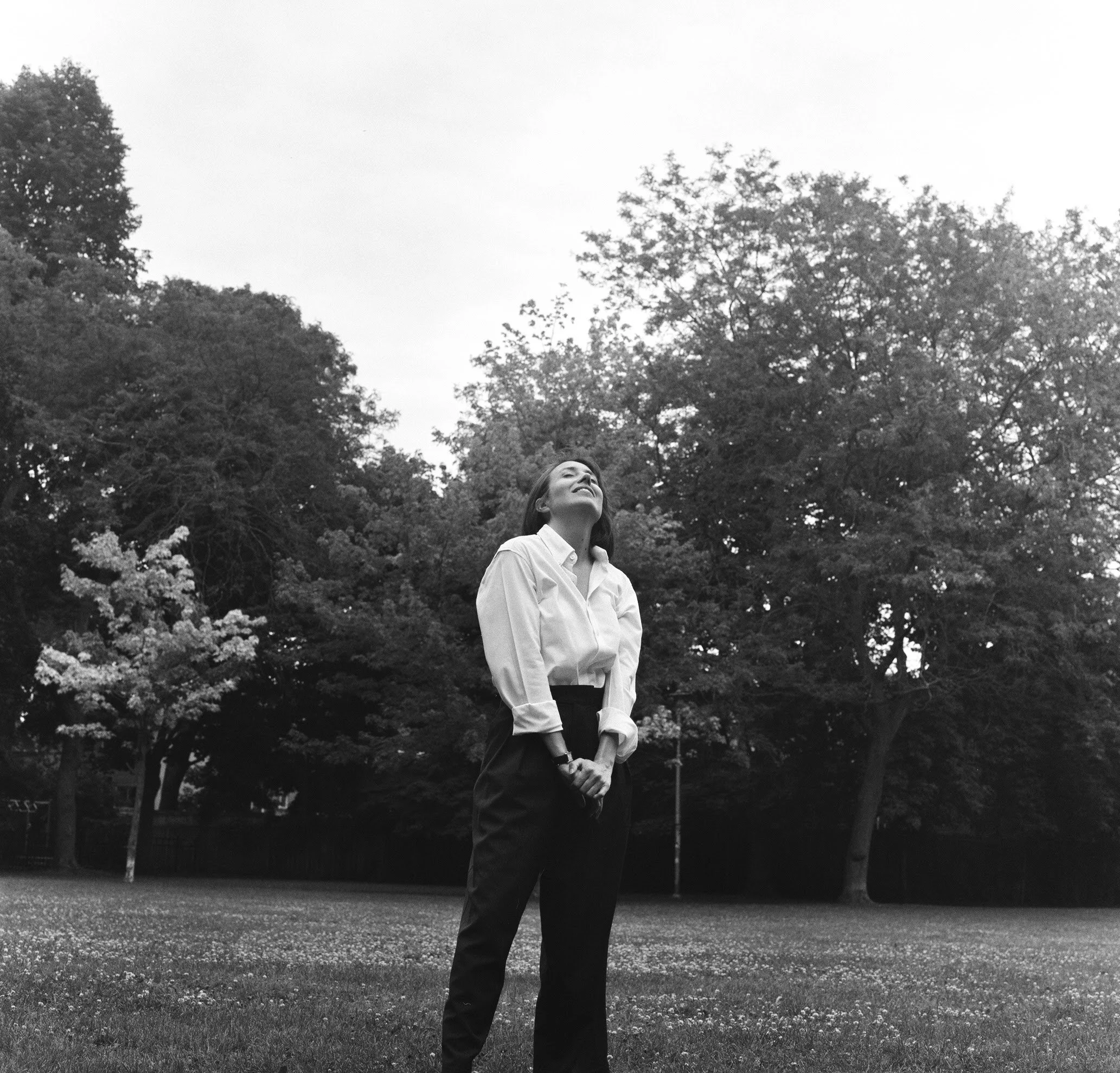 Mujer con vestimenta blanca y pantalones oscuros de pie en un parque con árboles y flores, mirando hacia arriba con expresión de alegría.