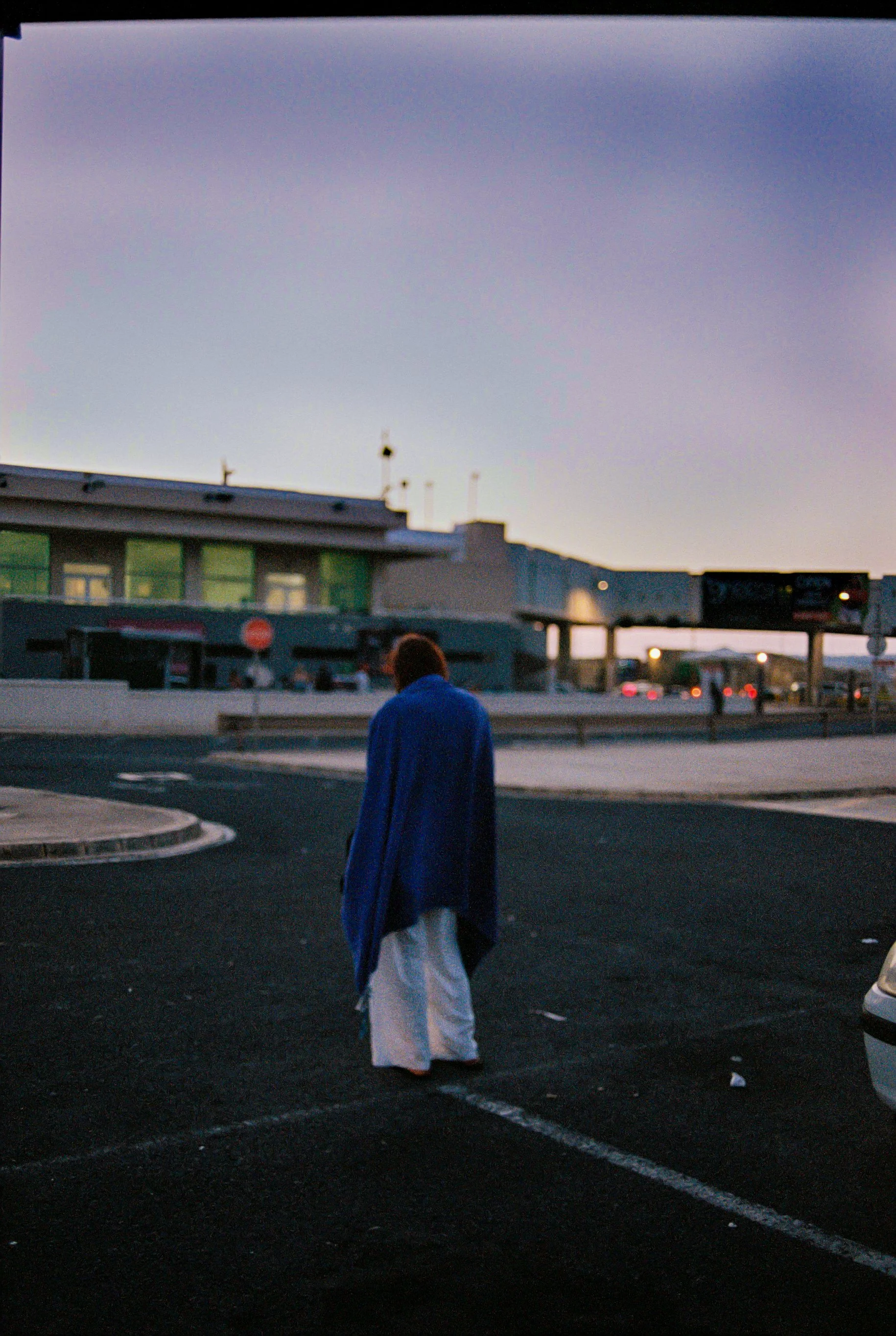 Persona con vestimenta tradicional caminando en un estacionamiento al atardecer, con edificio y cielo violeta al fondo.