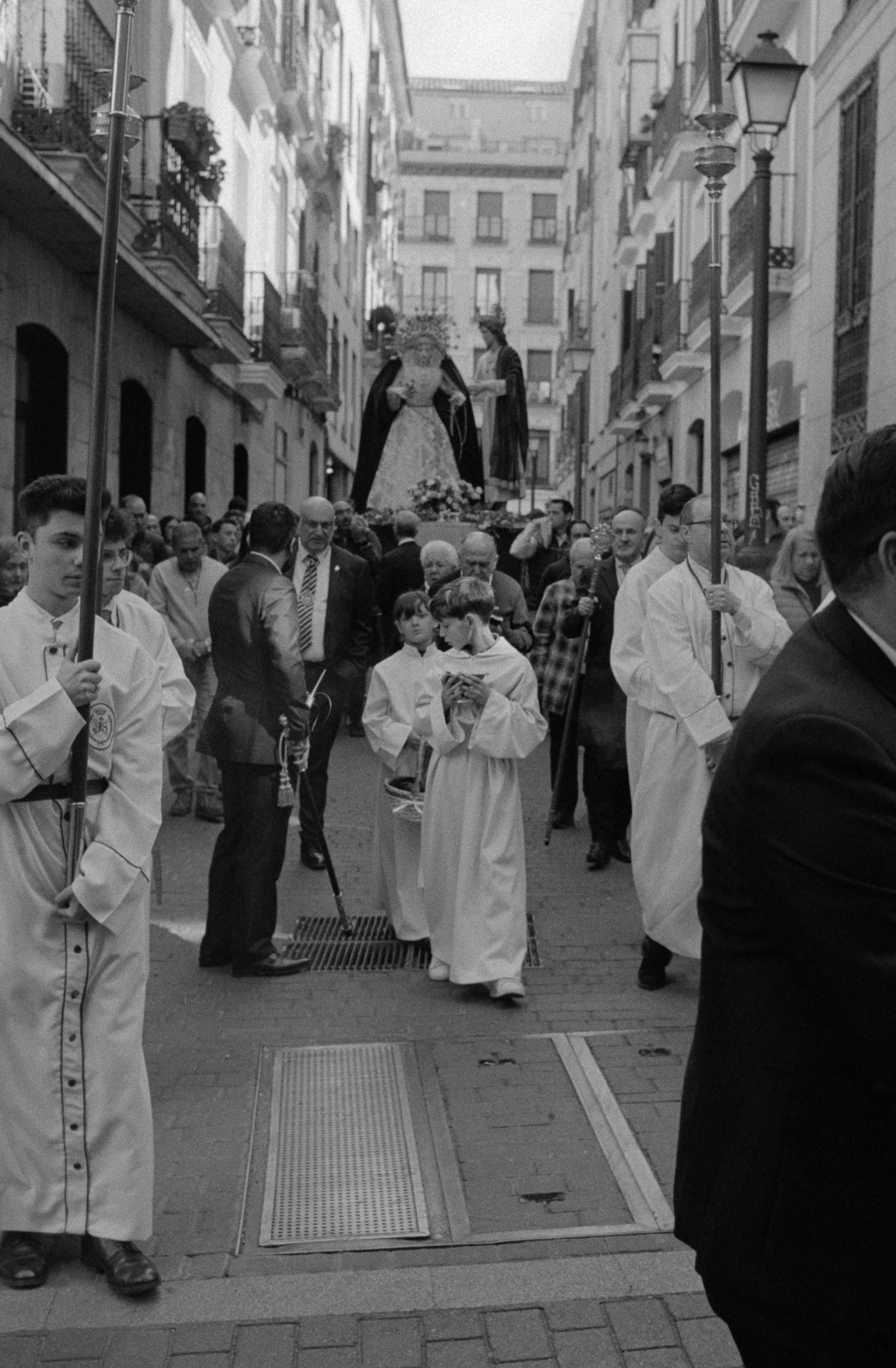 Una procesión religiosa con personas en calle estrecha, algunas con mantillas y vestimenta tradicional, llevando una imagen de Santa María. Muchas personas observan el evento.