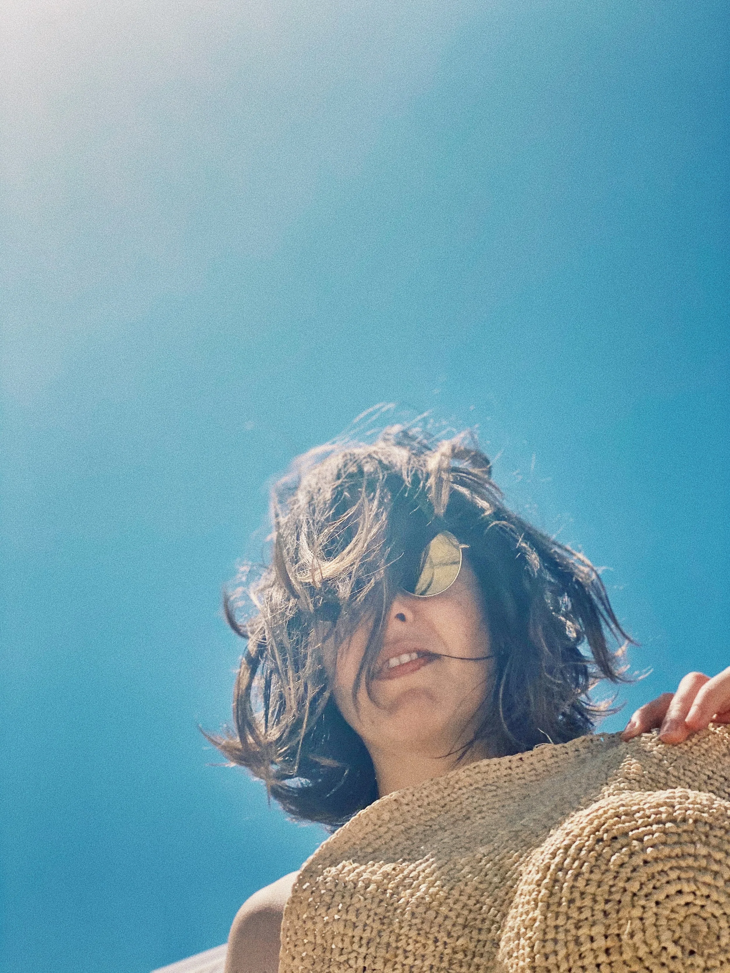 Mujer con gafas de sol y cabello rizado, usando un sombrero de paja, en un día soleado con cielo azul visible.