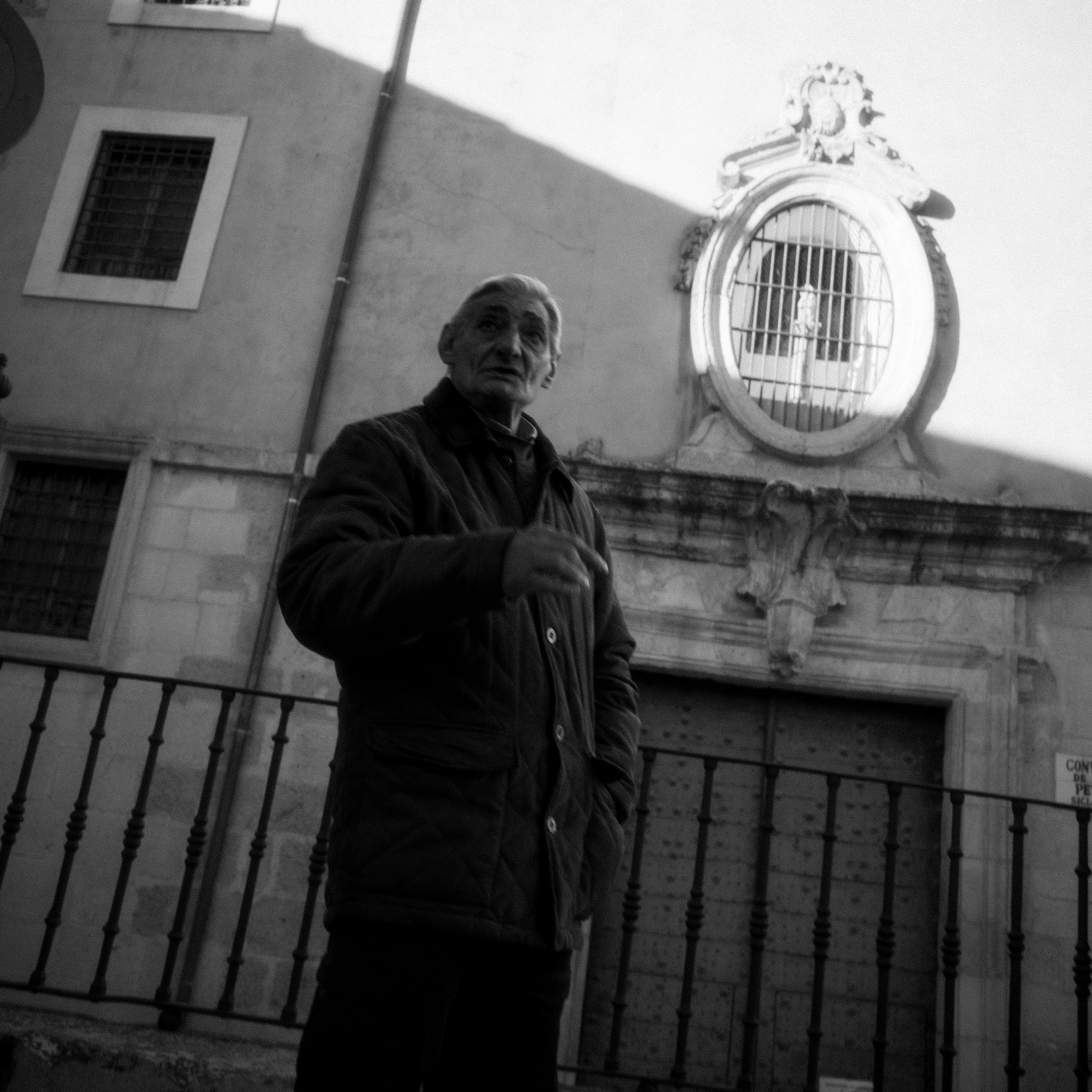 Hombre mayor con barba y cabello canoso, vestido con chaqueta oscura, de pie frente a una verja de metal en una calle de ciudad. Al fondo, un edificio con ventanas y una estructura decorativa en la parte superior.