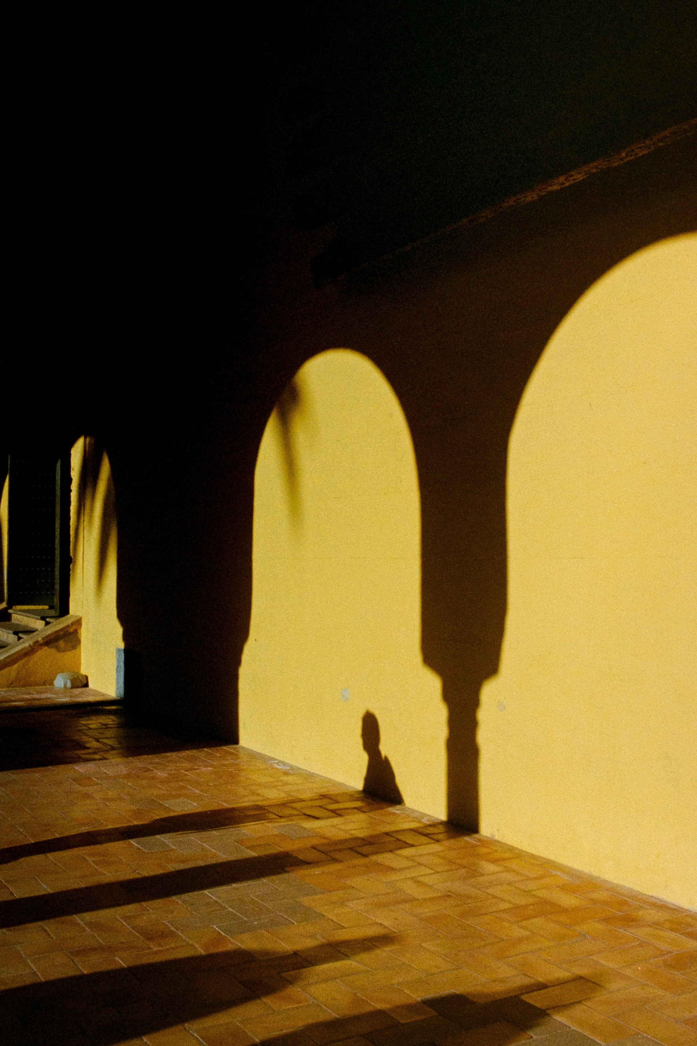 Sombras proyectadas de arcos en una pared amarilla, reflejadas en un suelo de baldosas de terracota, en un espacio interior con escaleras a la izquierda.