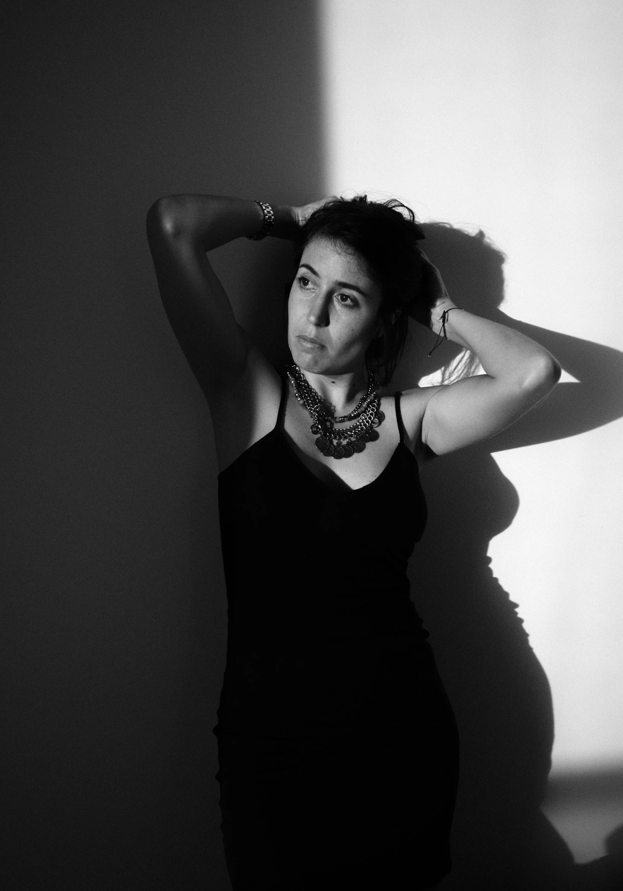 Retrato en negro y blanco de una mujer con expresión pensativa, con las manos en la cabeza y un colgante en el cuello, con sombra marcada a un lado de su cara.