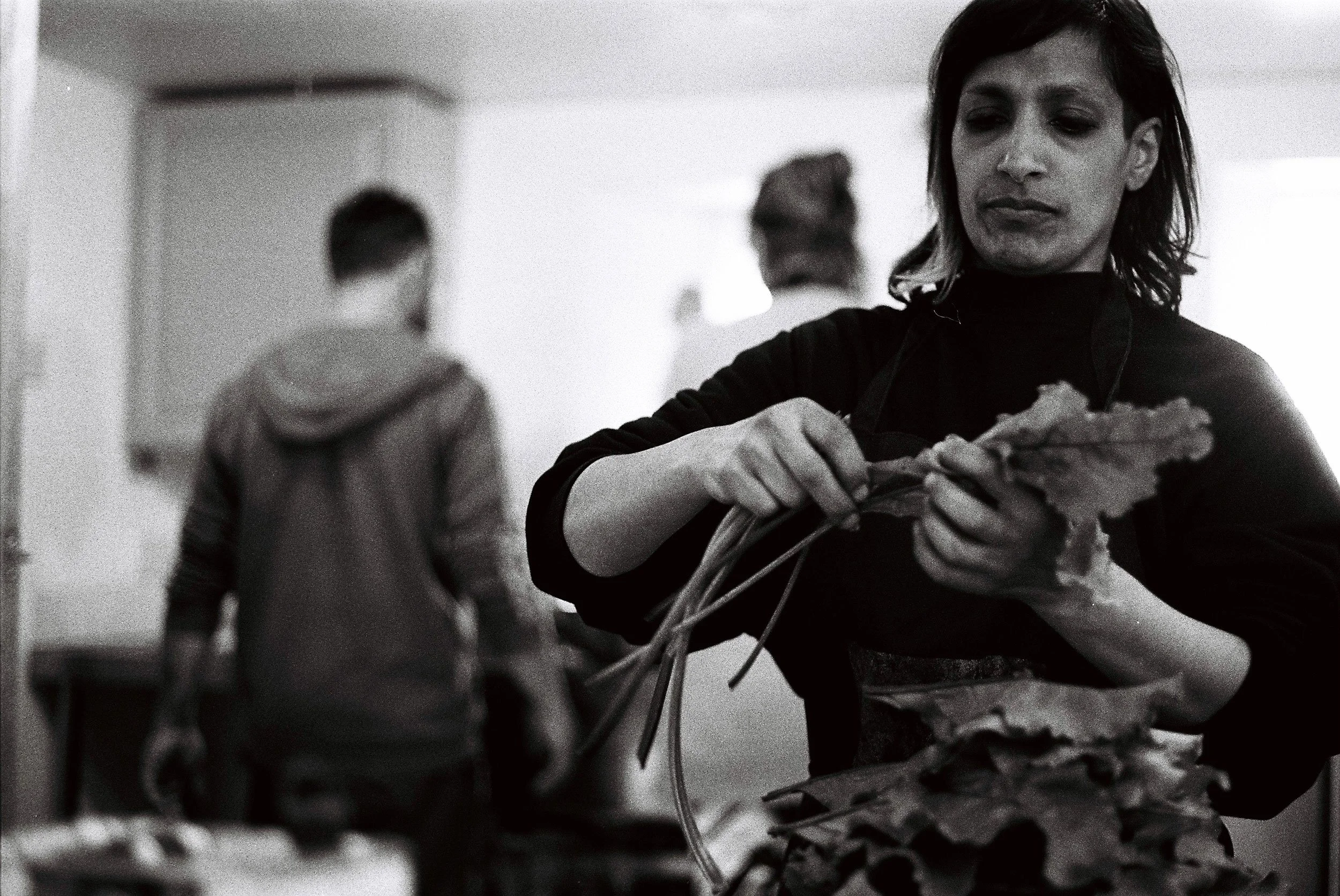 A woman in a black shirt and apron prepares leafy greens in a kitchen, with two people in the background, one wearing a hoodie and the other with glasses.